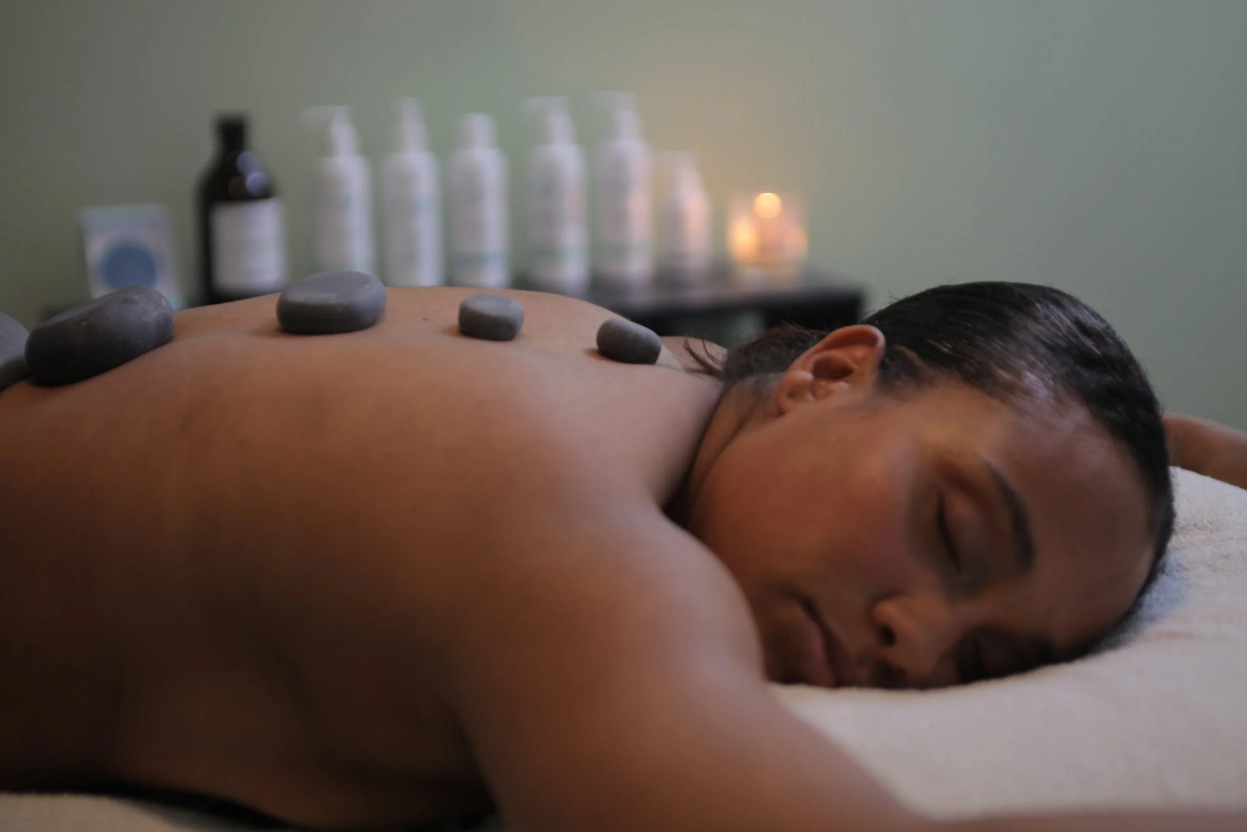 A person lying face down on a massage table with several smooth stones placed along their back, in a spa or massage setting.