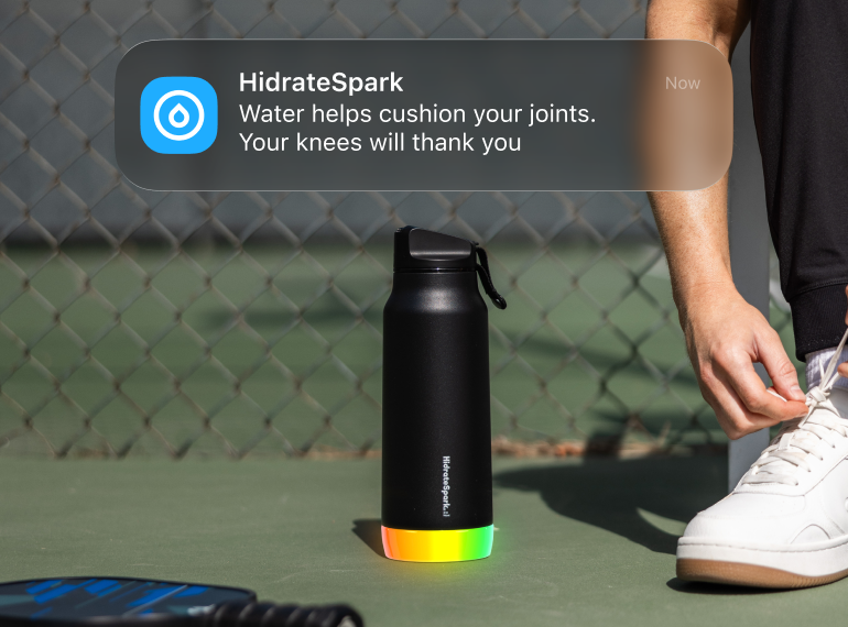 HydrateSpark smart water bottle glowing on a tennis court beside a person tying a white sneaker, with a hydration reminder notification overlay.
