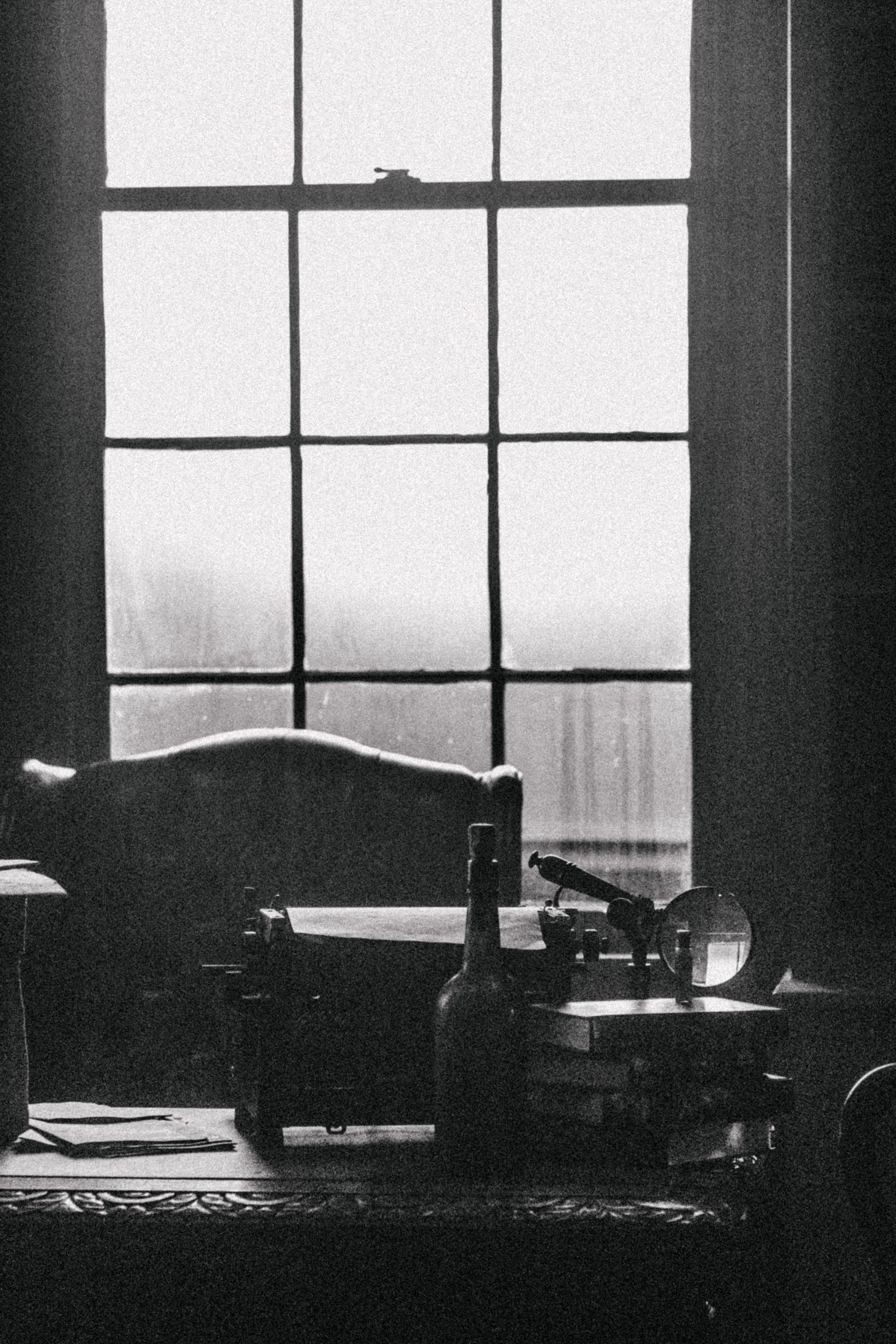 A black and white photo of a window with multiple panes, a table with various objects on it including a torch, small bottles, and glasses, and a decorative rug partially visible at the bottom.