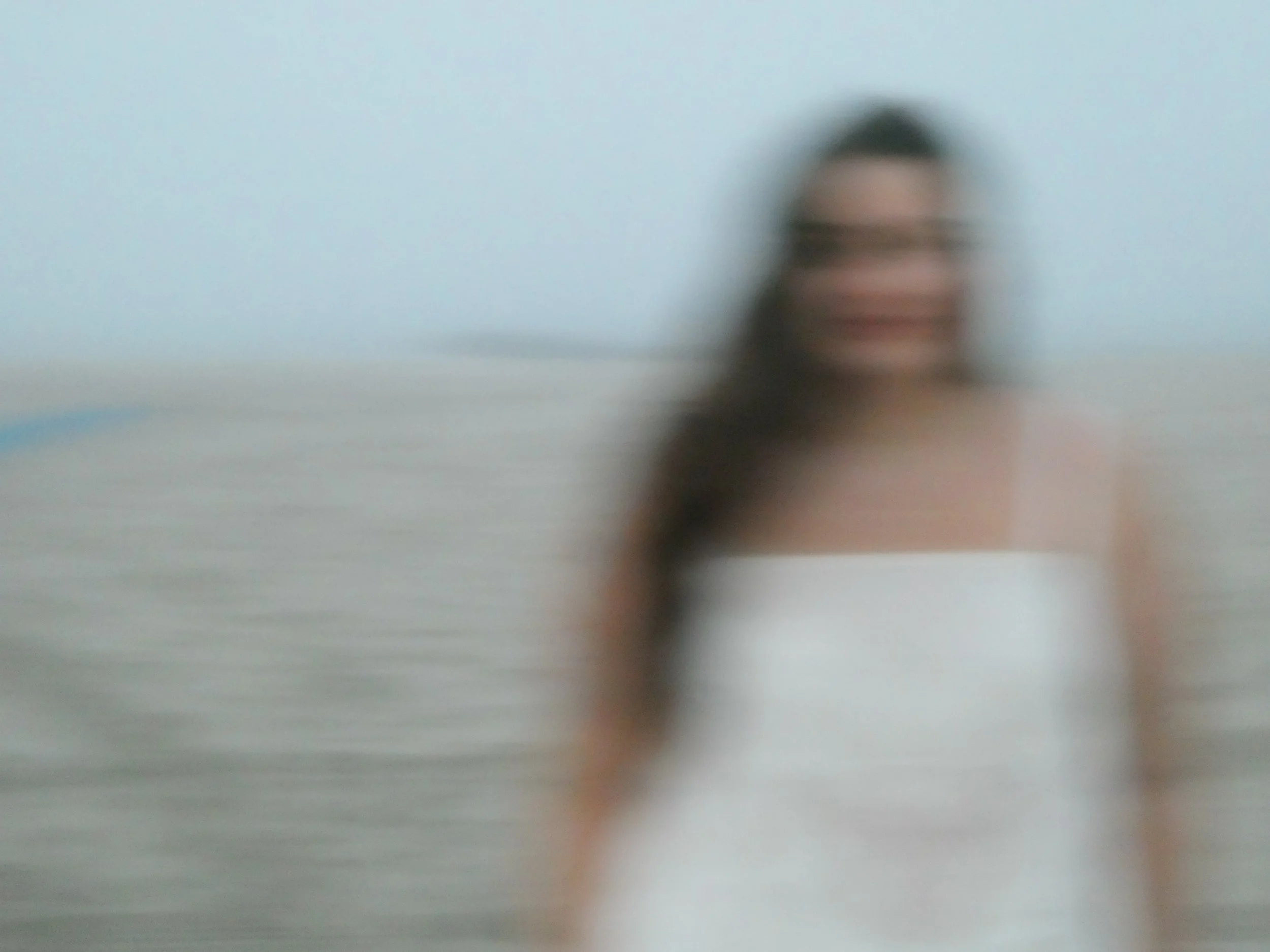 Blurred image of a person with dark hair, smiling, in front of a light blue background.