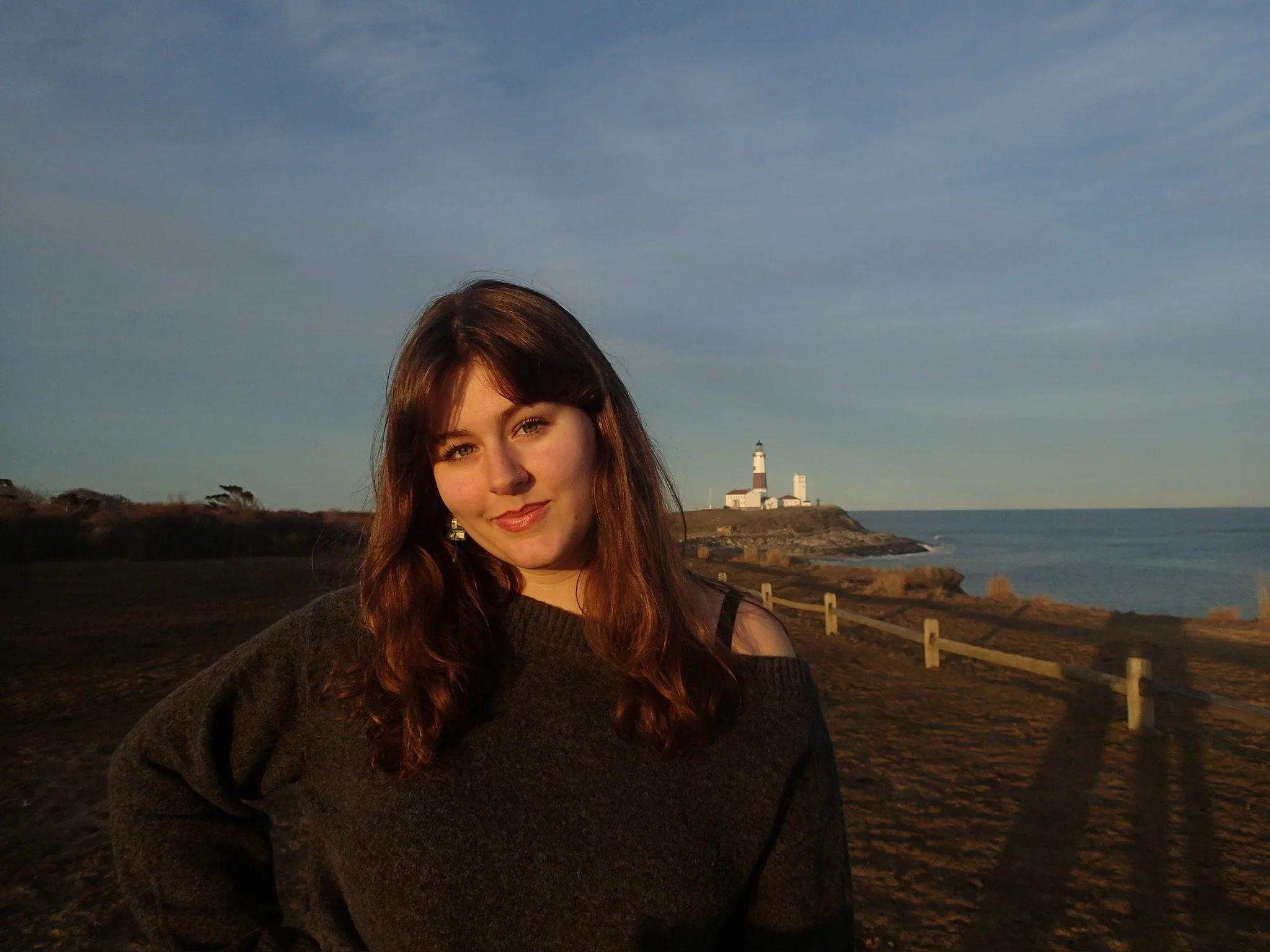 Young woman with long brown hair and a nose piercing standing outdoors near a coastline with a lighthouse in the background during sunset.