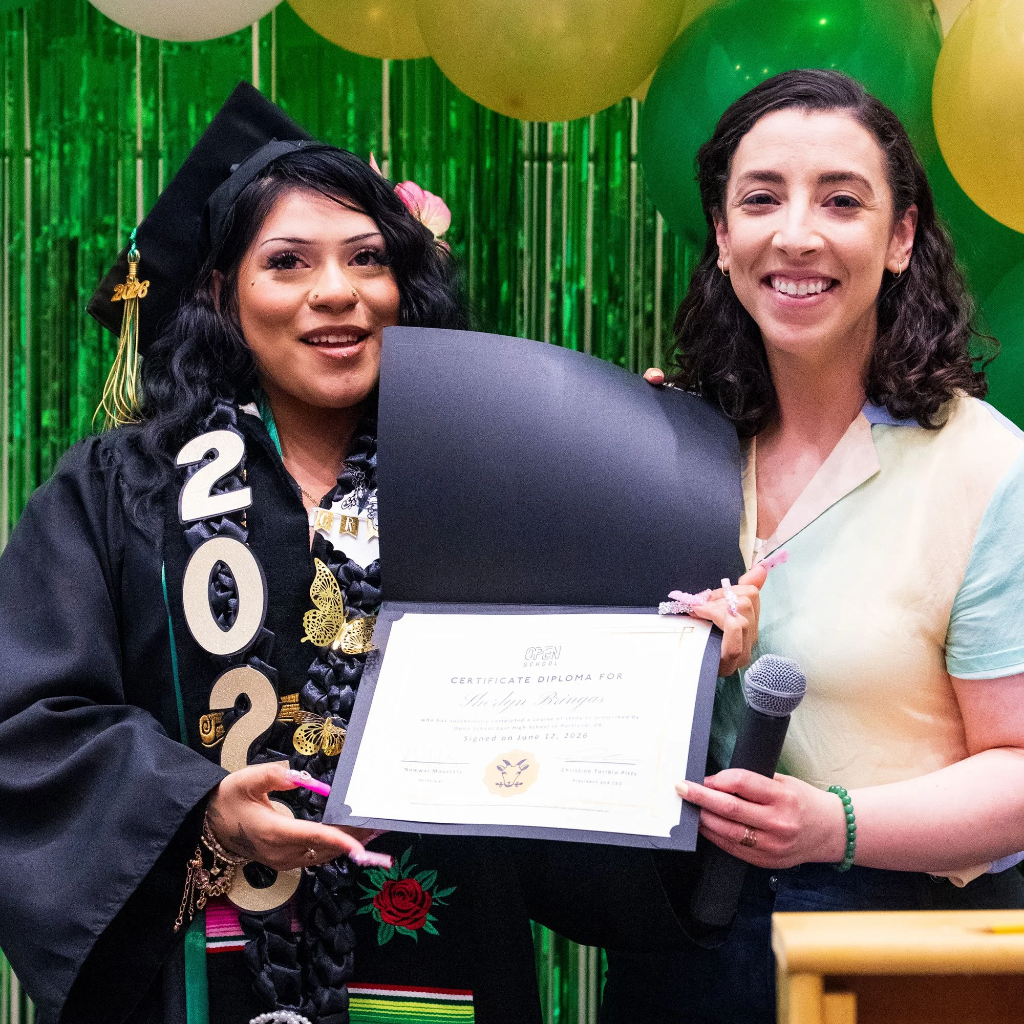 An Open School East graduate in cap and gown holding her diploma next to a smiling staff member holding a microphone