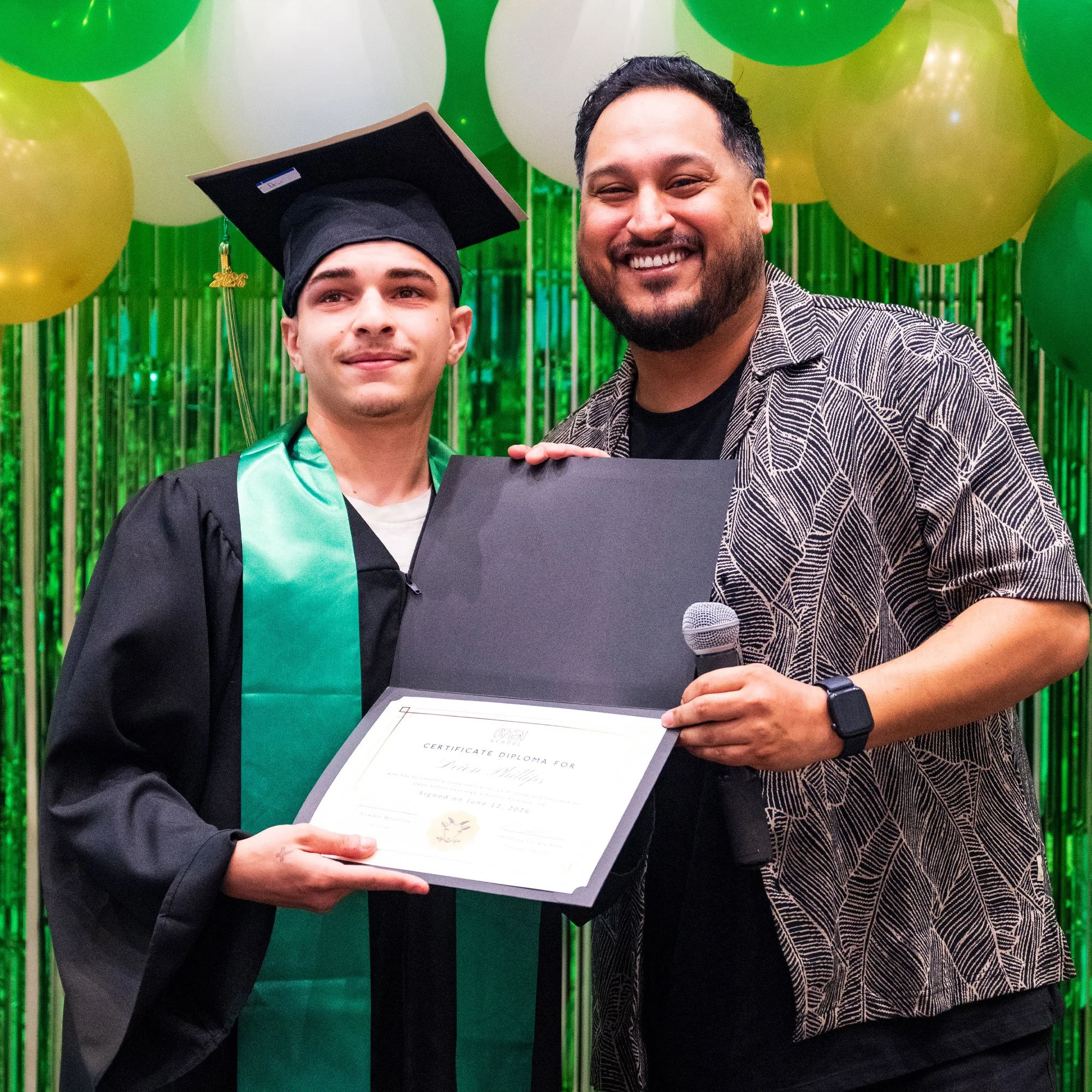 An Open School East graduate in cap and gown holding a diploma beside a smiling staff member