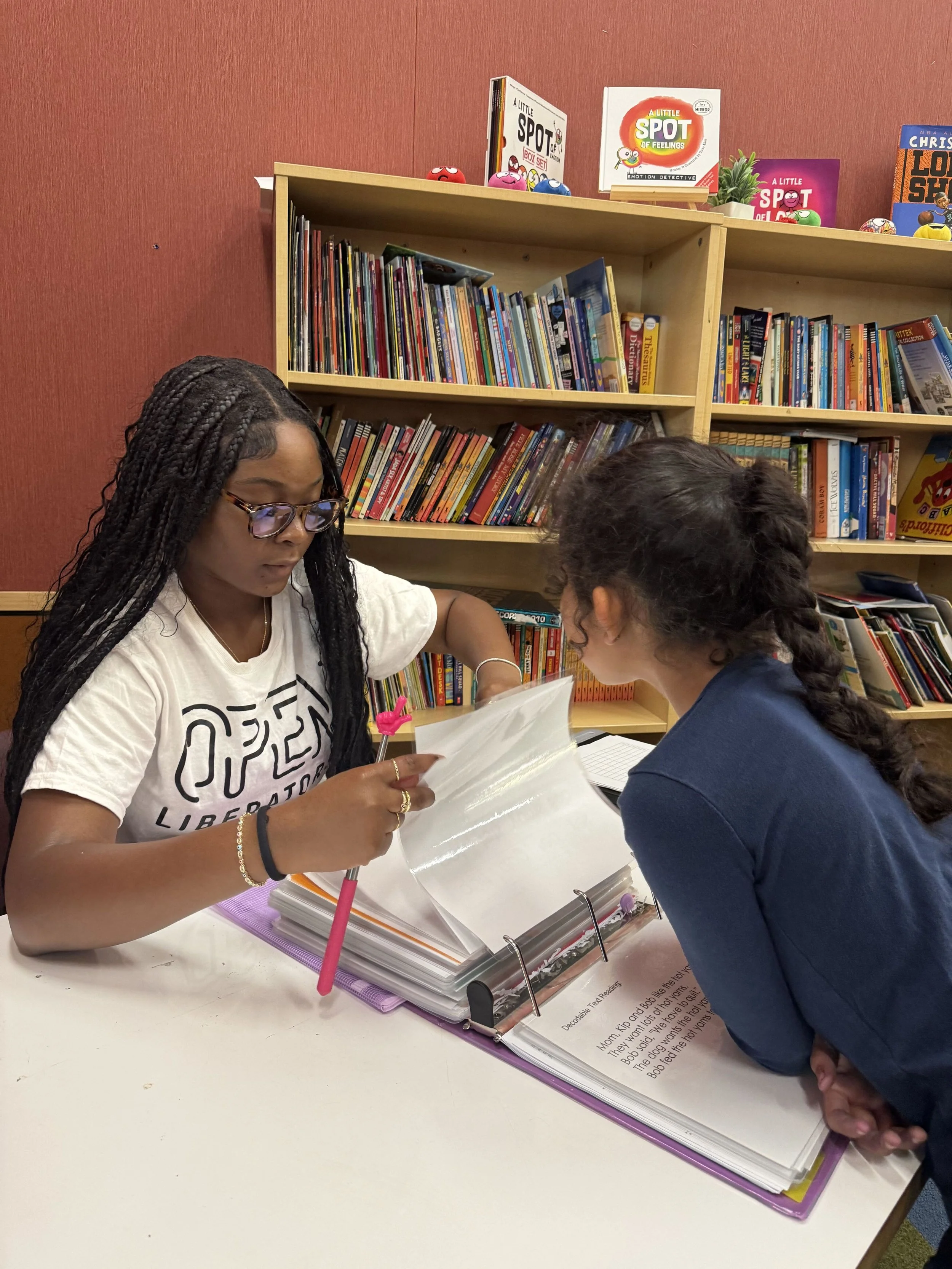 A Literacy Liberators tutor and a young student reading together from a book, with a bookshelf behind them