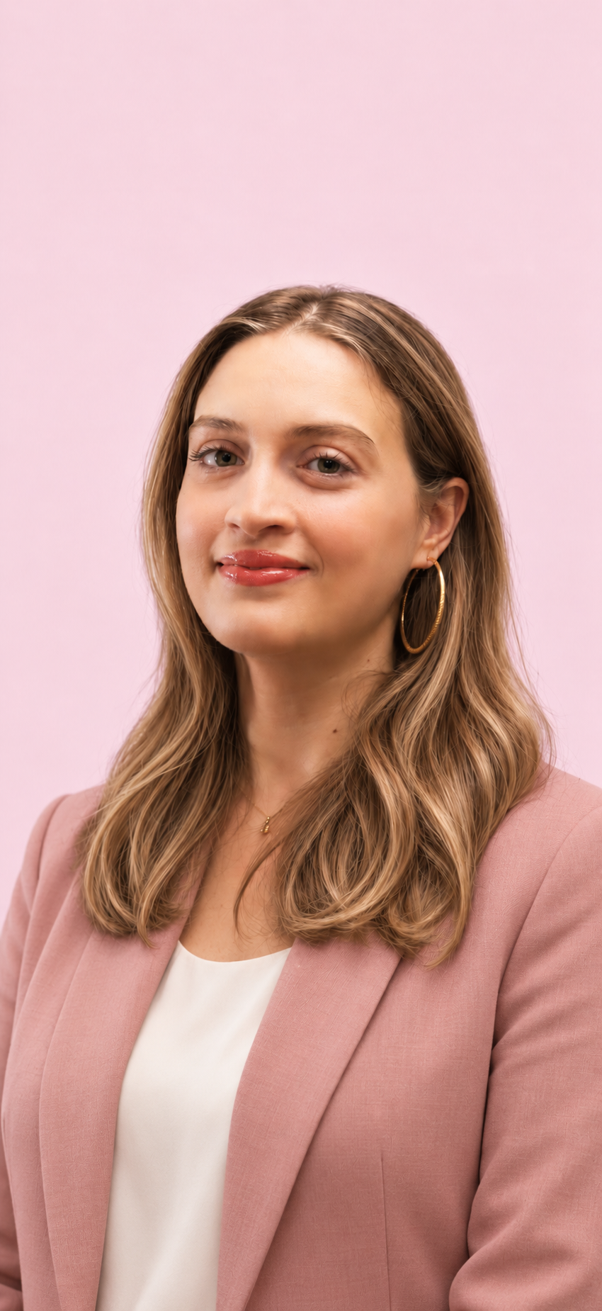 Une femme avec des cheveux longs et ondulés, portant un blazer rose et une chemise blanche, avec des boucles d'oreilles en or, se tenant contre un fond rose clair.