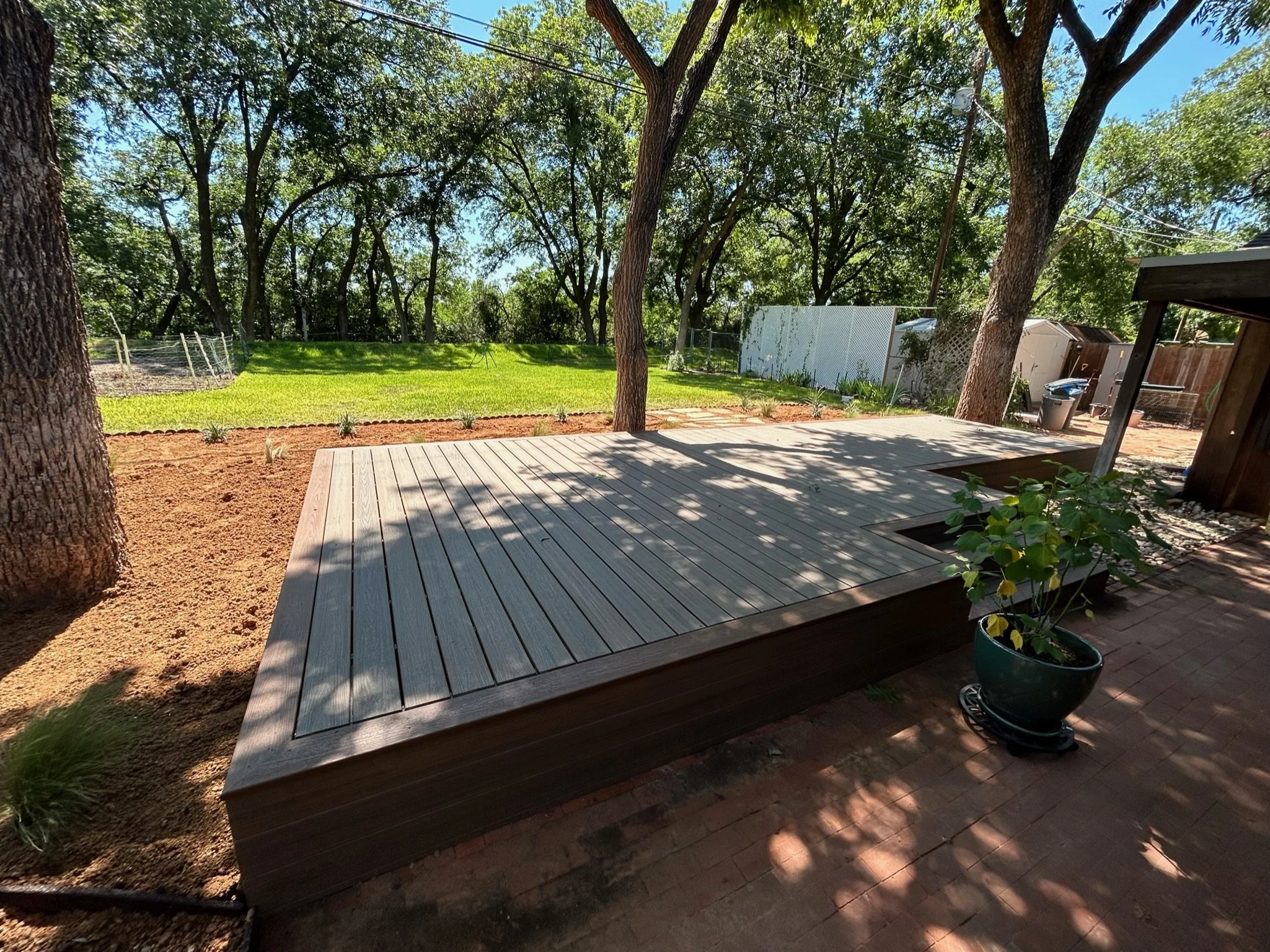 A backyard scene with a wooden deck, green grass, trees, and a fence, with blue sky visible through the tree leaves.