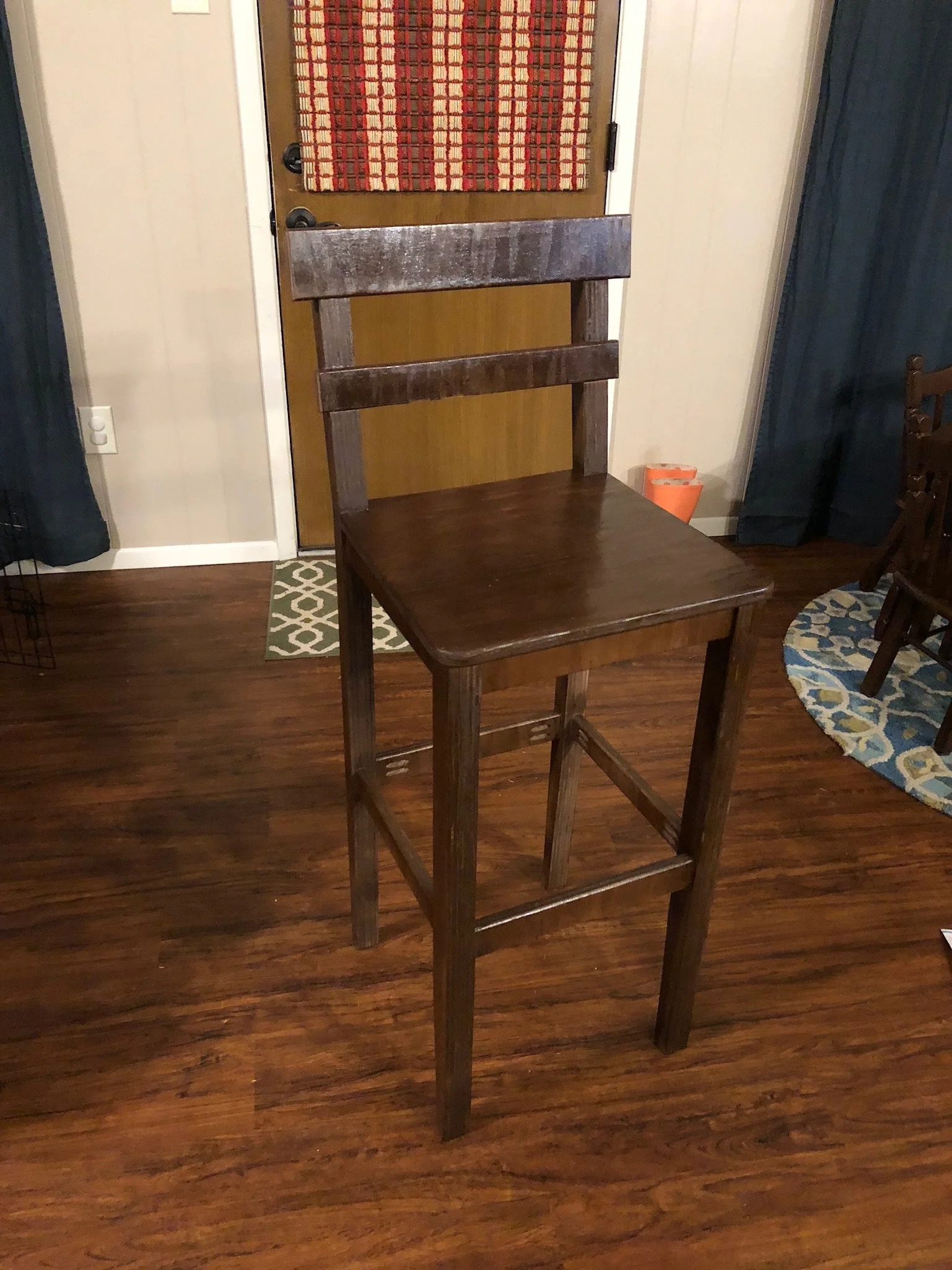 Unfinished wooden chair in a room with dark hardwood floors, a partially visible blue curtain, a small patterned rug, and a door with a red and beige striped decoration hanging on it.