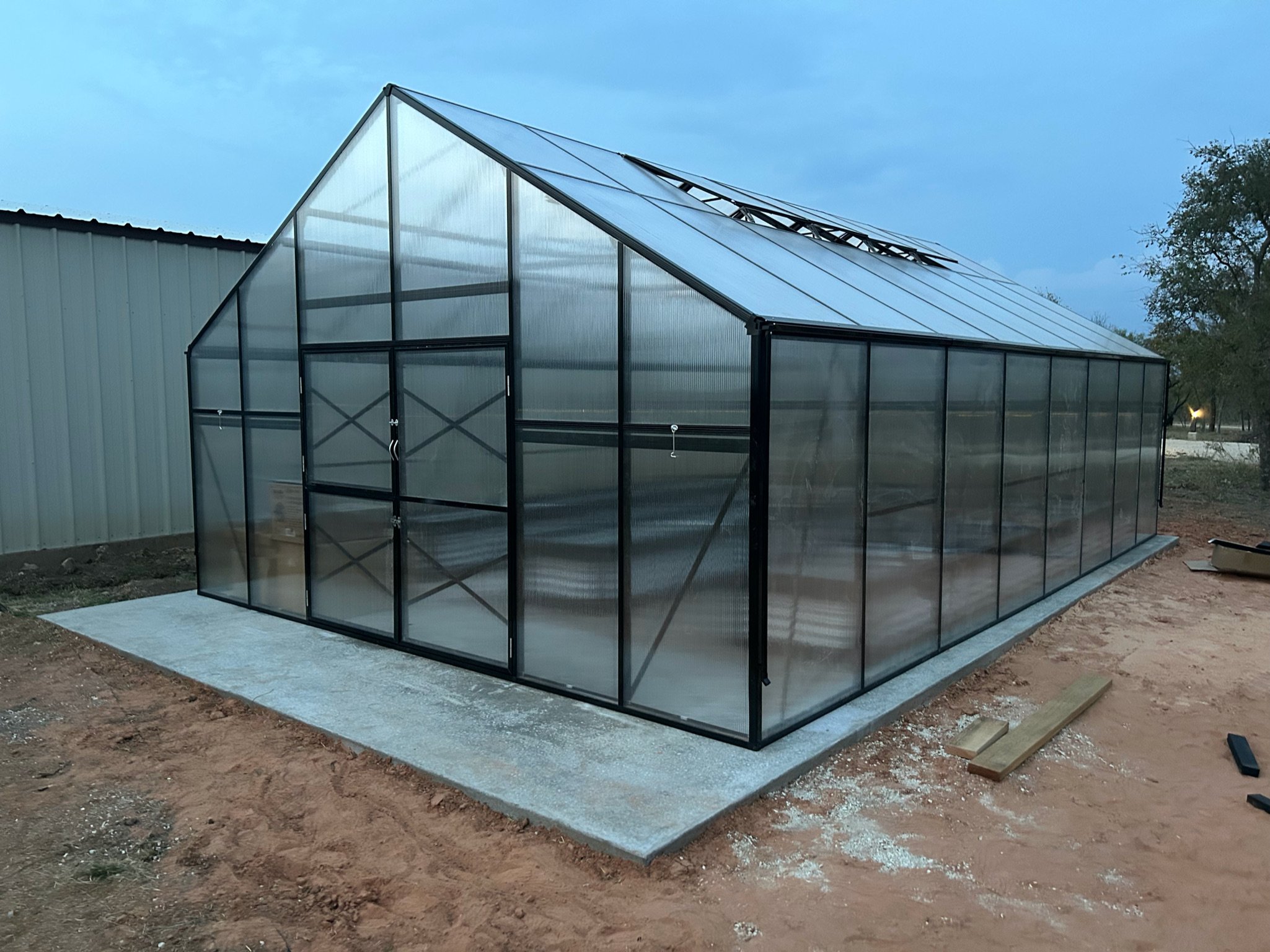 A metal and glass greenhouse structure on a concrete slab outside, with a cloudy sky and trees in the background.