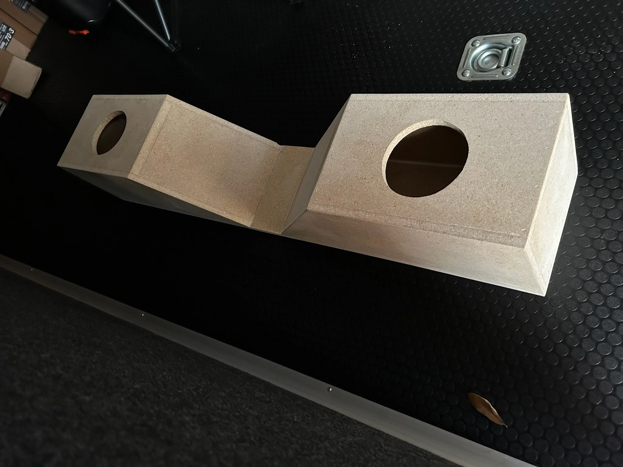 A wooden speaker box with two circular openings, placed on a black textured surface with a metal speaker mount.
