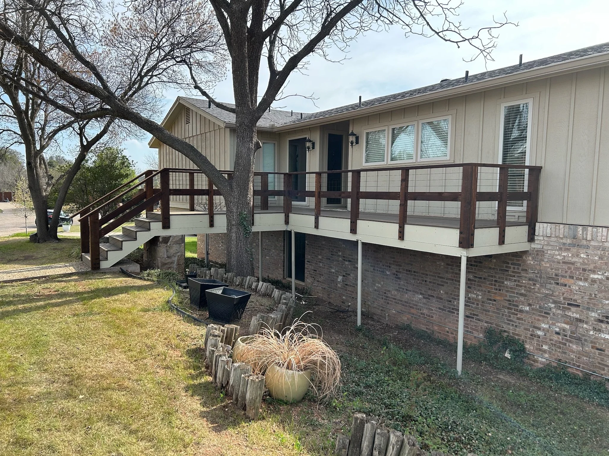 A two-story house with beige siding, multiple windows with white blinds, and a wooden deck with stairs leading to the yard. A tree with bare branches is in front of the house, with a small garden bed and planters underneath.