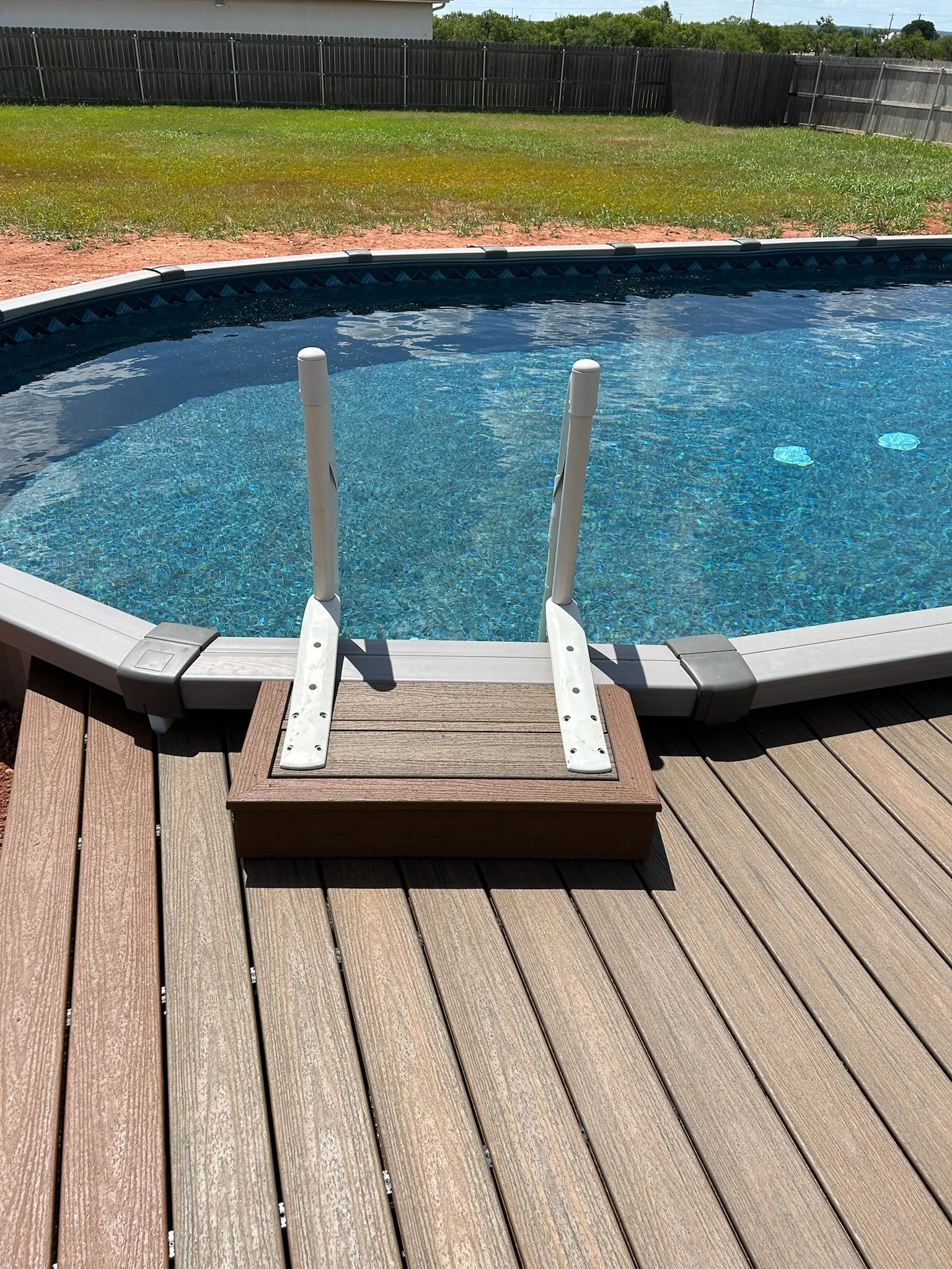 Backyard with a swimming pool and a wooden deck, surrounded by a fence and grassy area.