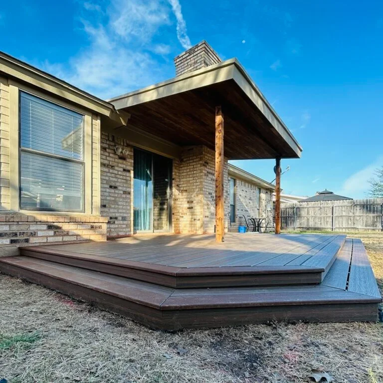 Backyard view of a house with a wooden deck and covered patio, fenced yard, and blue sky.