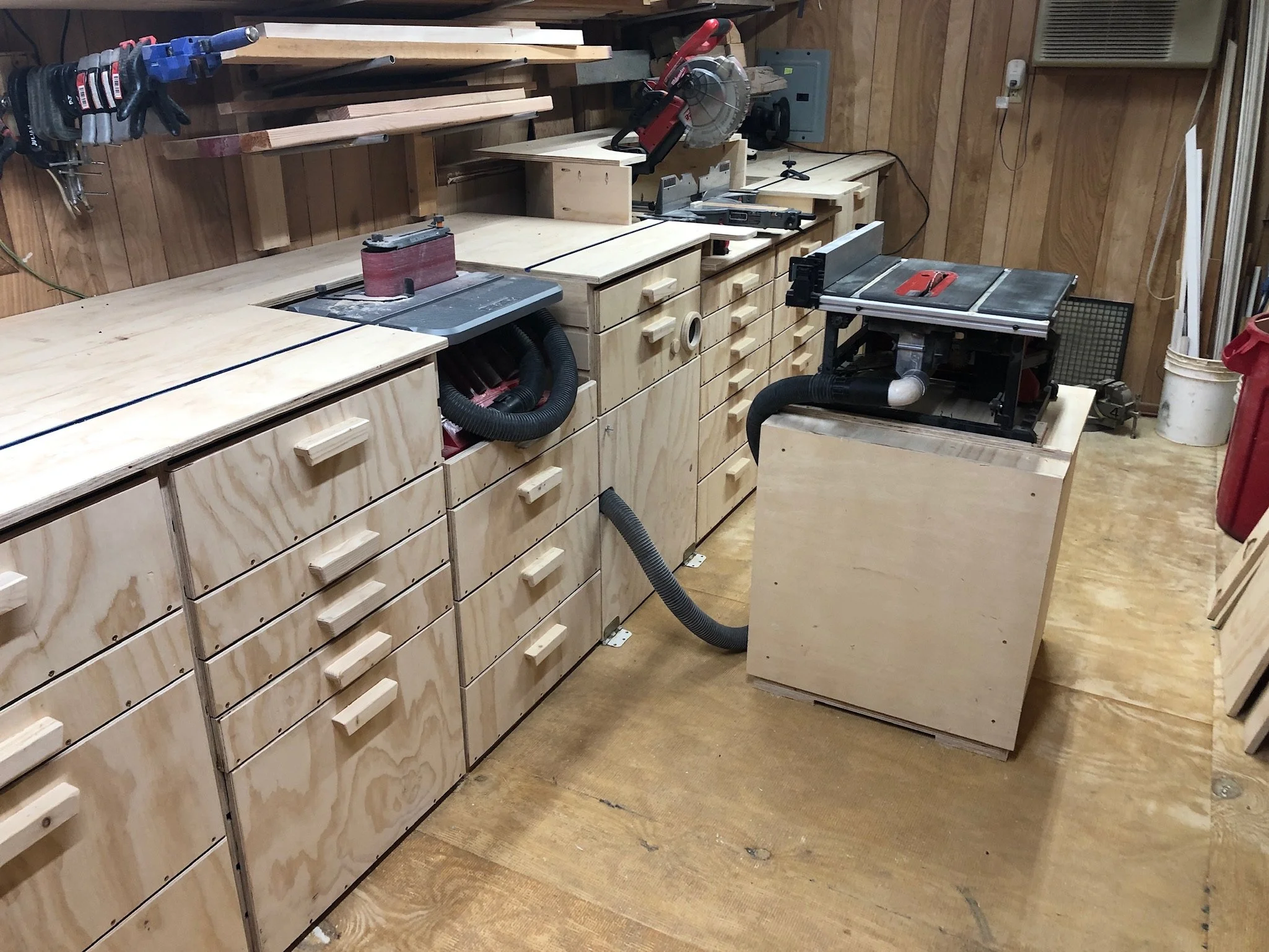 Woodworking workshop with multiple workbenches, tools, and storage drawers, against wooden paneled walls.