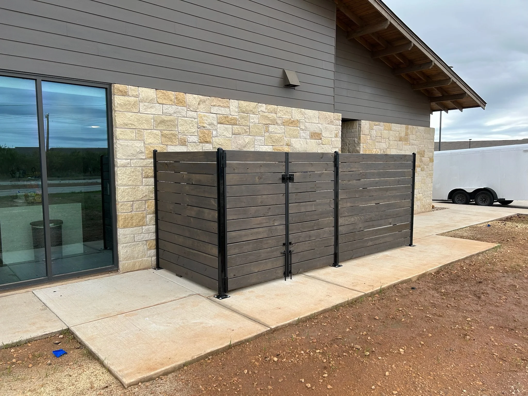 Side view of a modern house with gray siding and beige stone exterior, a large sliding glass door, a wooden privacy fence, a trailer, and a cloudy sky.