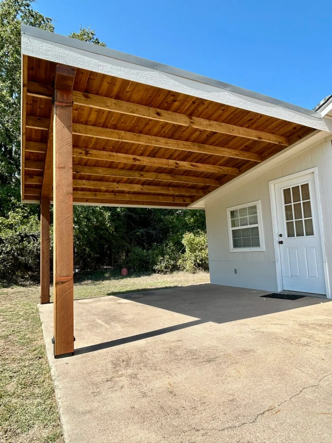 Newly built wooden covered patio attached to a white house with windows and a door, on a concrete slab with a grassy yard and trees in the background, under a blue sky.