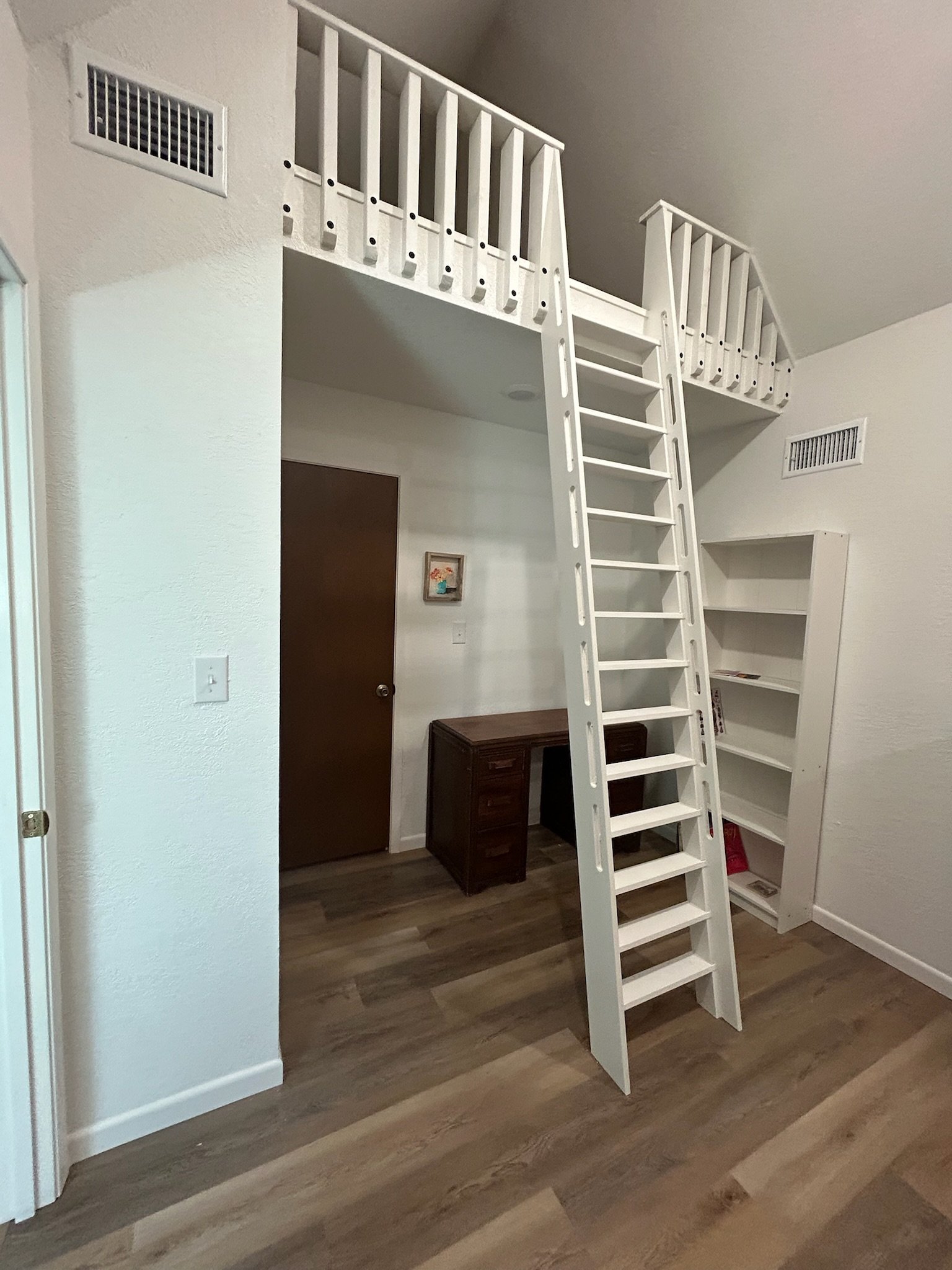 A white loft bed with a ladder is installed in a room, with a white bookshelf positioned next to it. The room has white walls, dark wood flooring, a small framed picture, and a brown door.