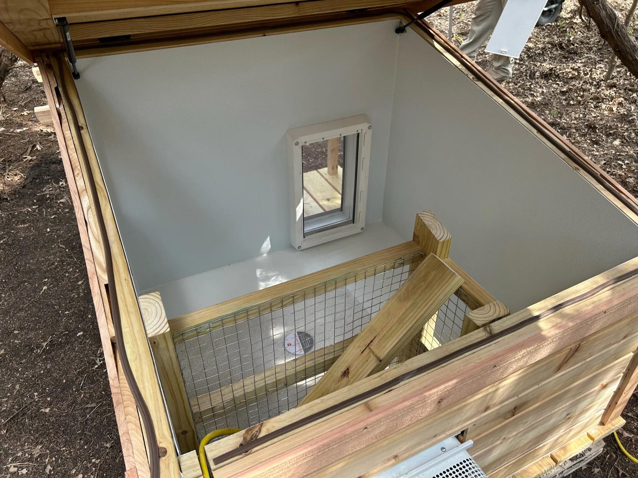 Wooden framed small structure with white interior walls and a small window, outdoors on dirt ground.