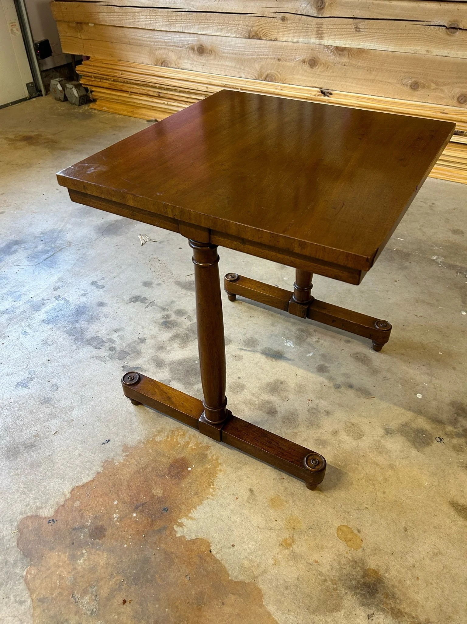 A wooden table with a dark finish on a concrete floor in a workshop or garage. The table has turned legs and a rectangular top. There are wooden planks leaning against a wall in the background.