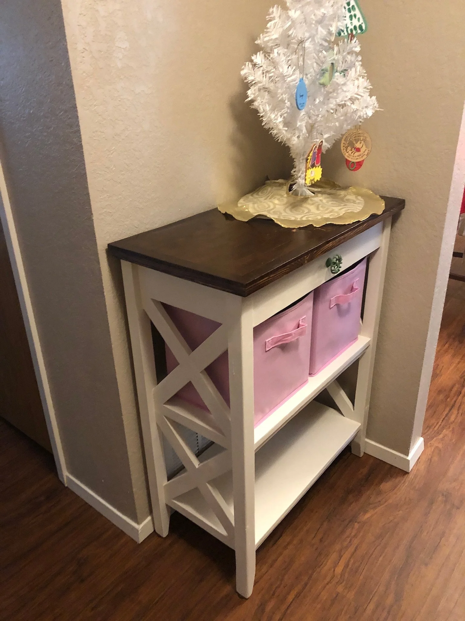 A small white decorative Christmas tree with ornaments, sitting on a green and gold tray, placed on a white and dark wooden shelf unit.