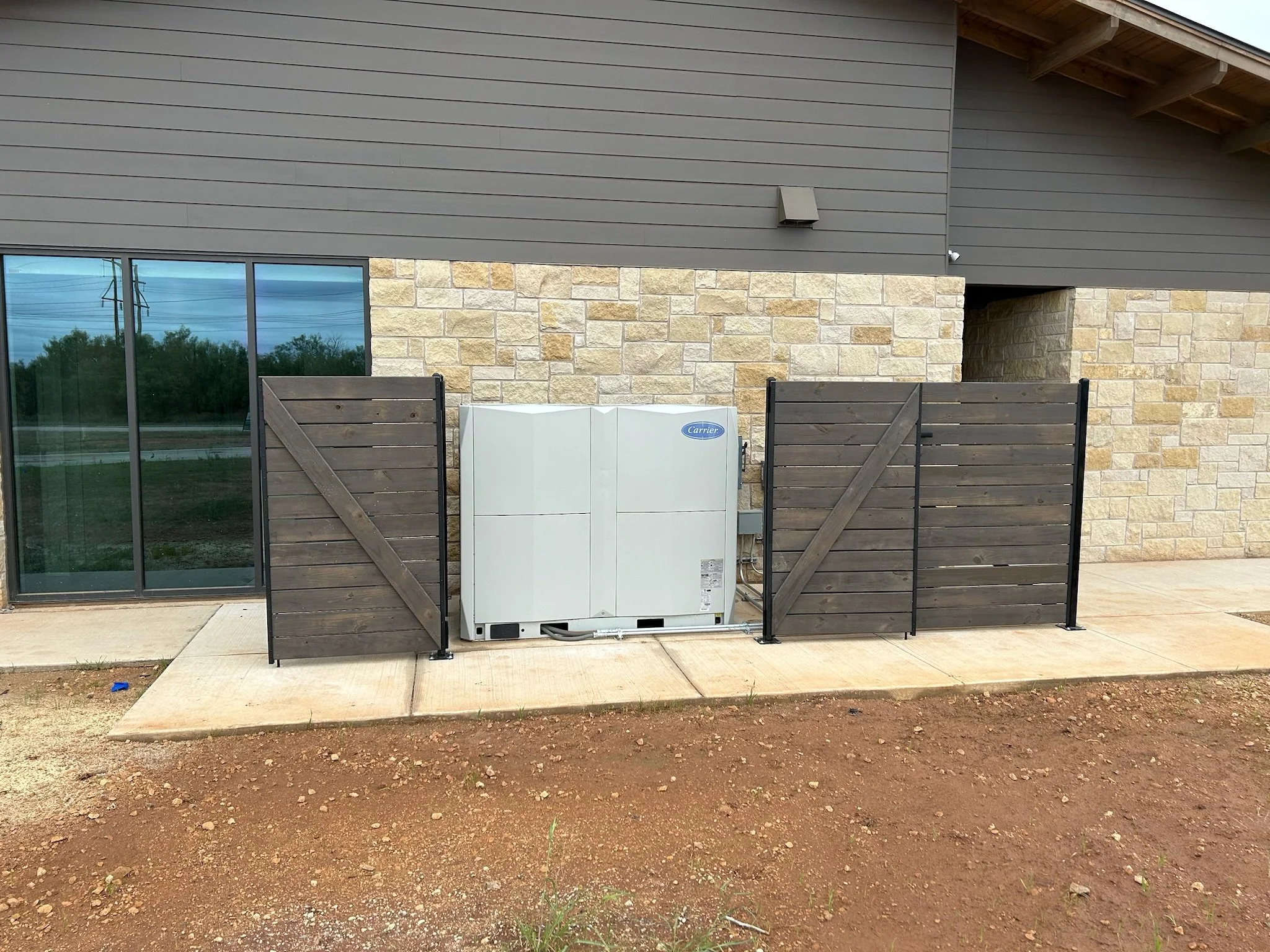 Exterior view of a building with stone and gray siding, a window, and HVAC unit on the ground, surrounded by a wooden fence gate and open dirt and concrete sidewalk area.