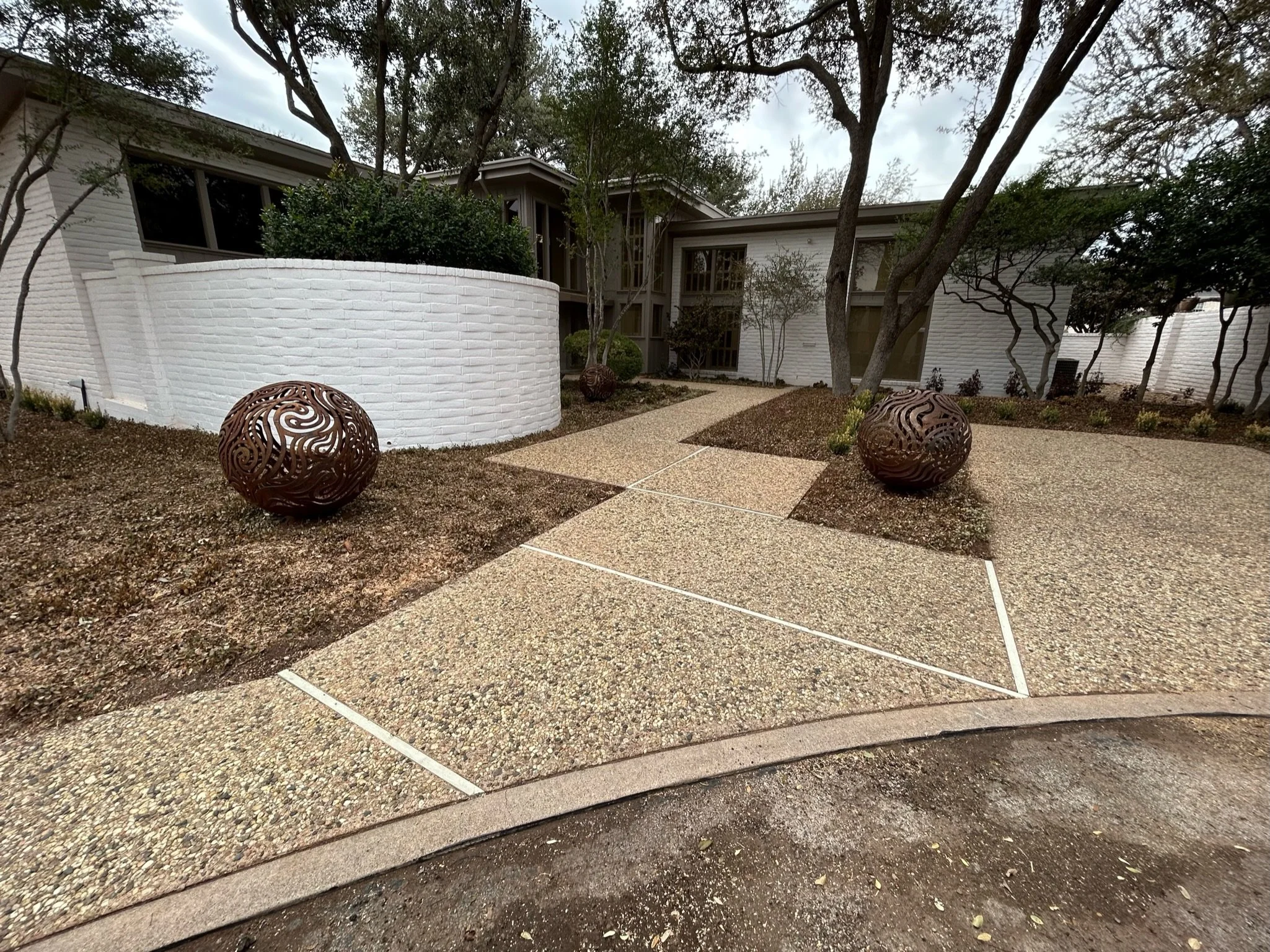 Modern house with white brick exterior, tall windows, surrounded by trees and landscaped garden with decorative spheres, gravel pathways, and concrete slabs.