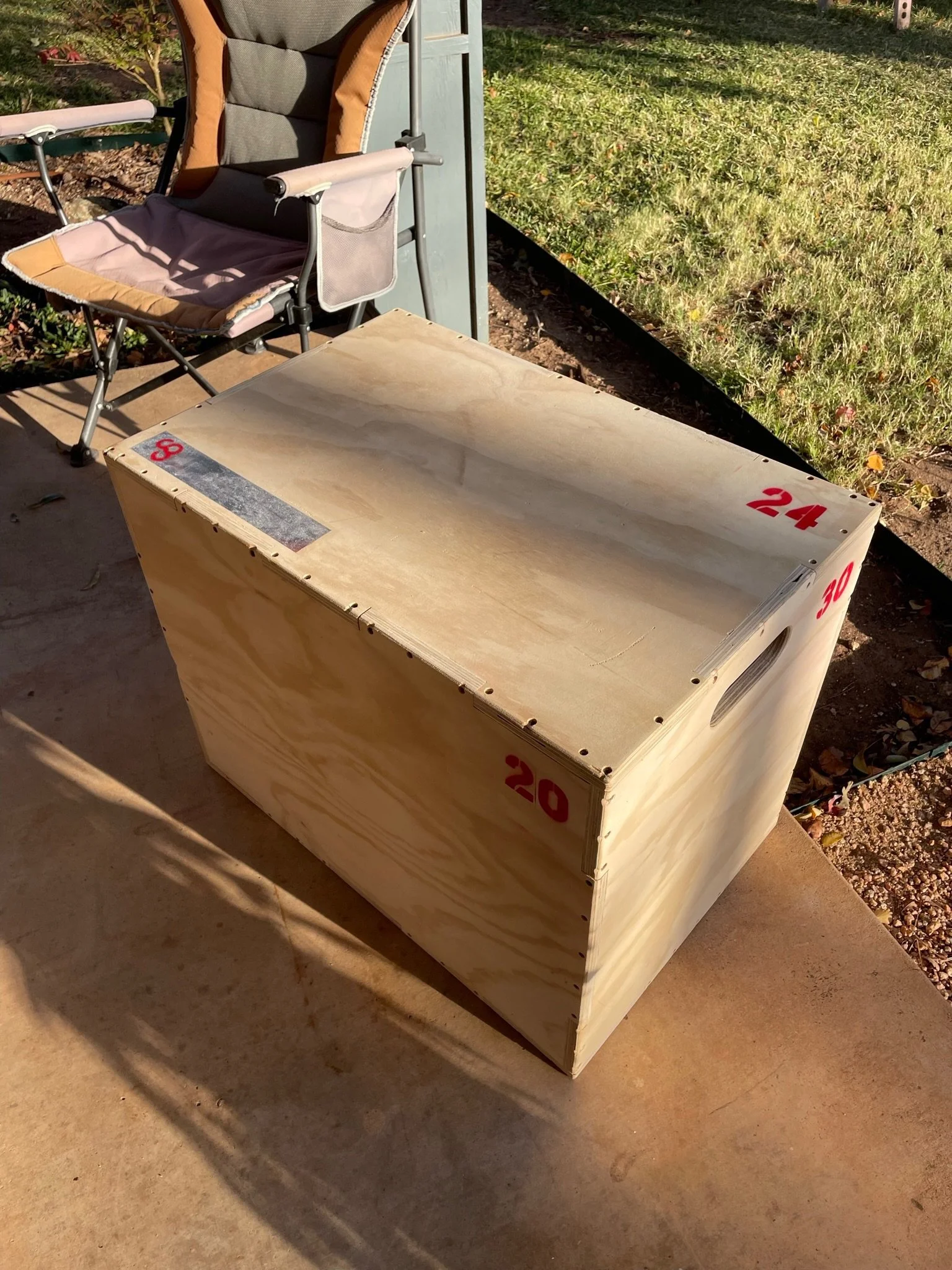 A wooden box with red numbers 20, 24, 28, and 30 printed on its sides, positioned on a concrete surface outdoors. Behind it, there is a folding chair with padded armrests and a dark, cushioned seat and backrest, next to a planter box with green plant