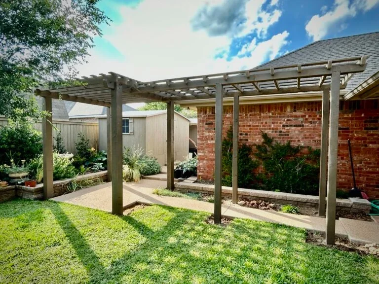 Backyard with a wooden pergola, garden beds, and a brick house wall.