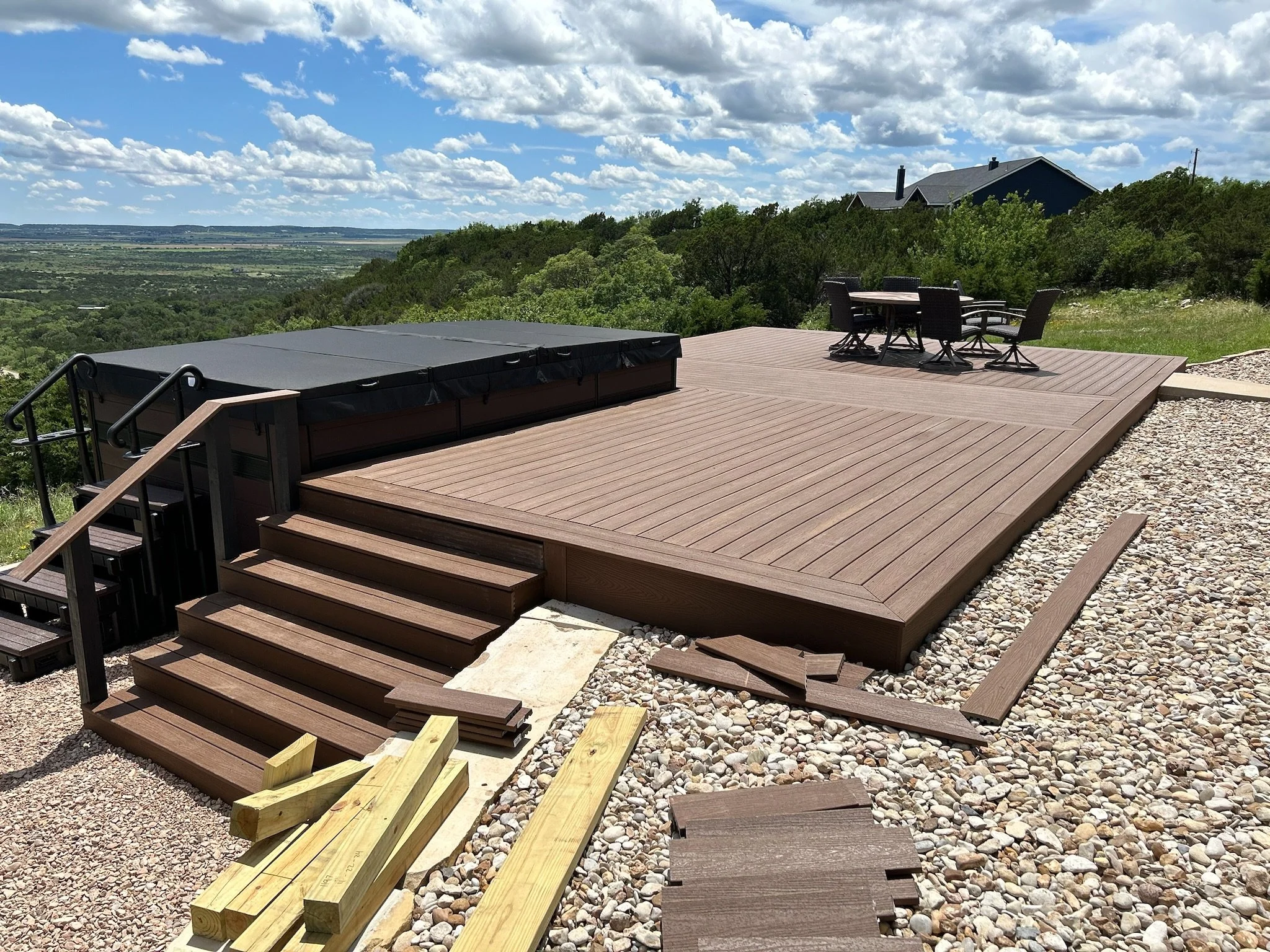 A backyard deck with a hot tub, outdoor table and four chairs, overlooking a scenic view of rolling green hills and a house in the distance under a partly cloudy sky.