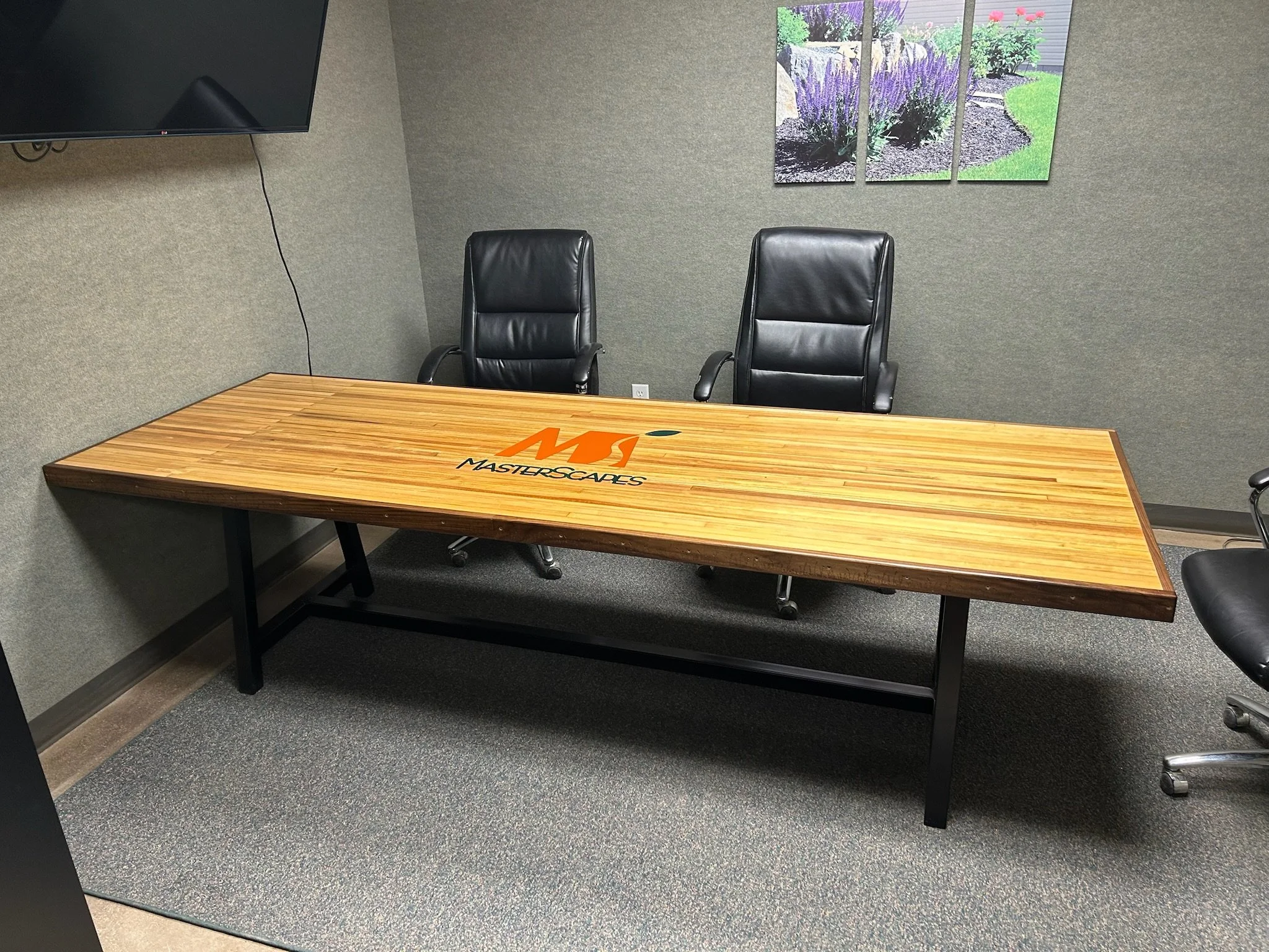 An office conference room with a wooden table and two black leather chairs, a large tv mounted on the wall, artwork of a garden scene on the wall, gray carpeted floor, and a power outlet on the wall.