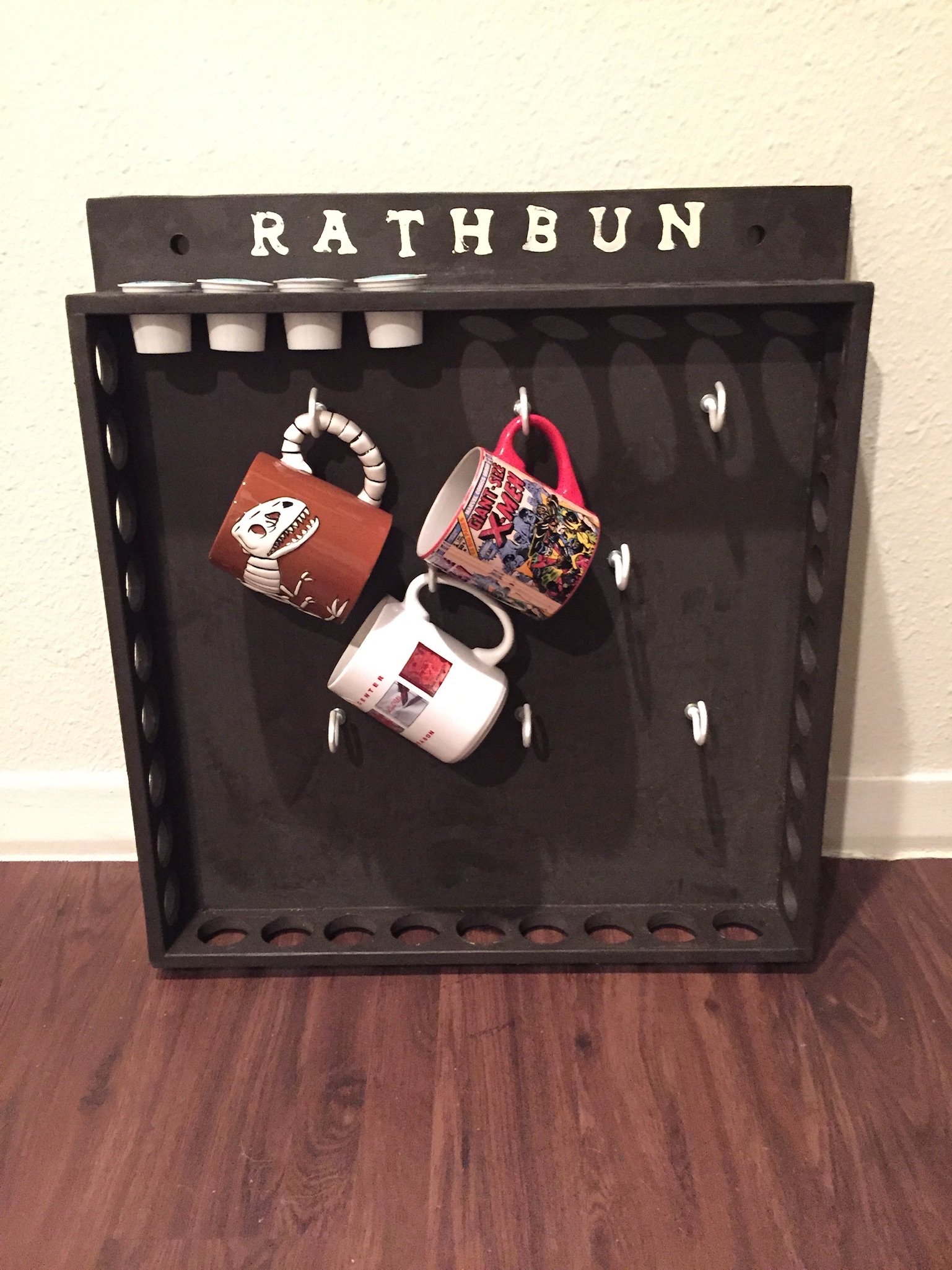 A black wall-mounted coat rack with the word 'RATHBUN' cut out at the top. The rack has four hooks, with two hanging mugs and a white plastic item.