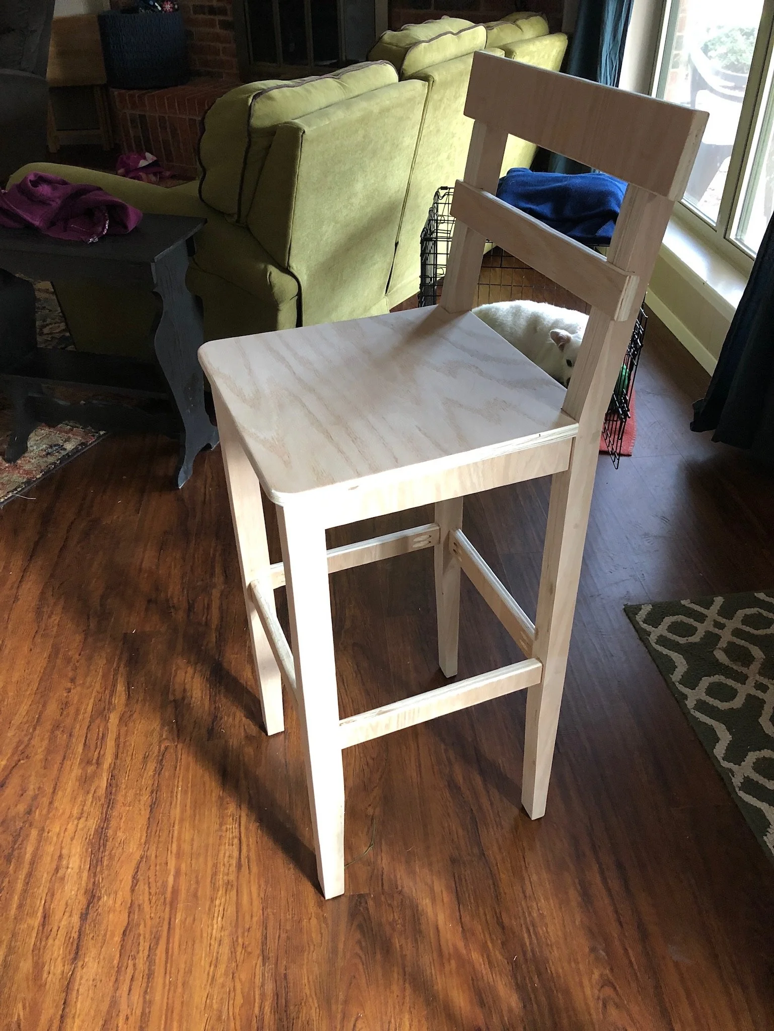 Unfinished wooden chair in a room with hardwood floors, with a yellow-green couch, a black side table, and a window with blue curtains in the background. There is a small dog lying in a wire crate behind the chair.