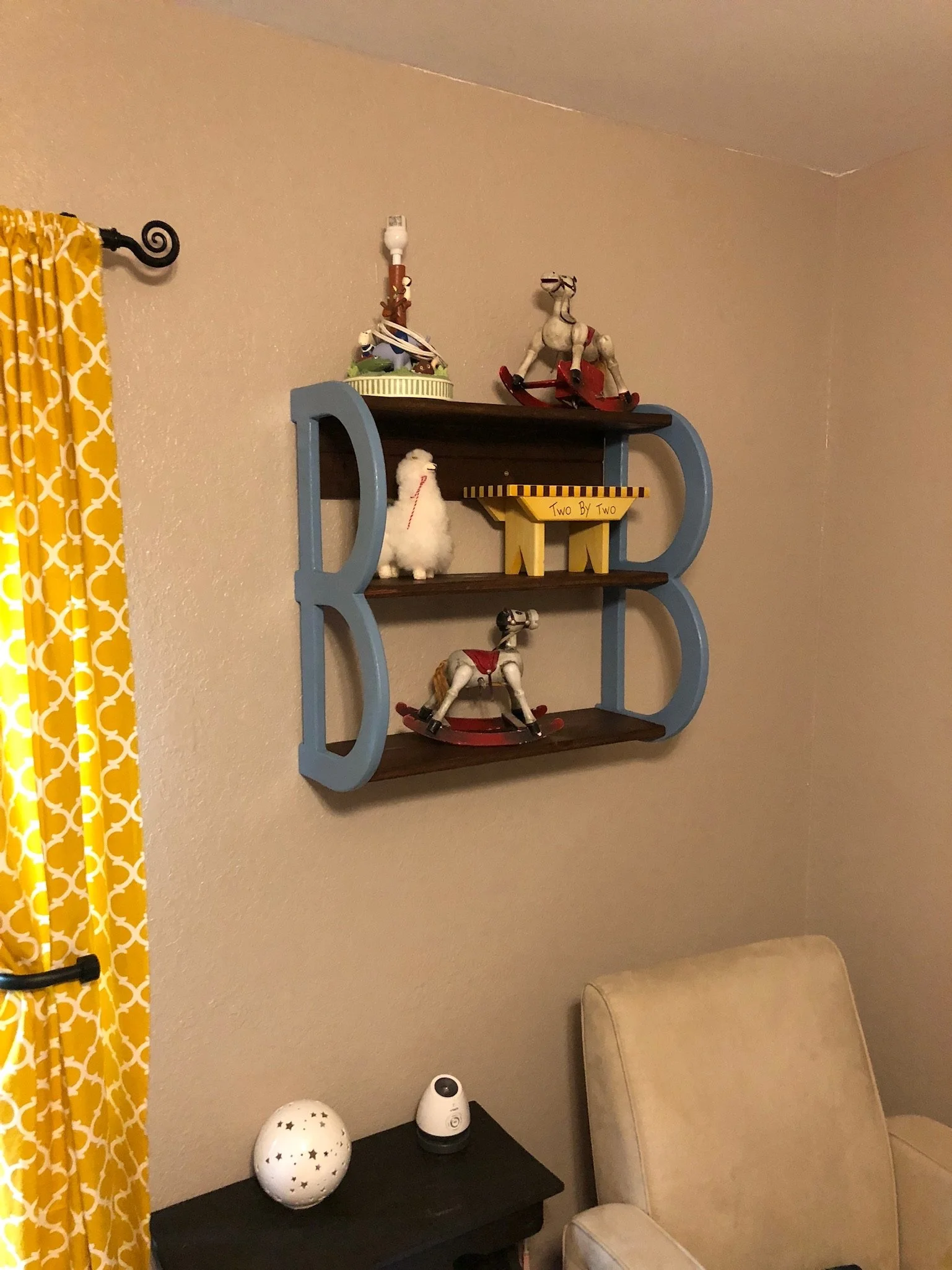 A blue wooden wall shelf with three levels decorated with Christmas-themed items, including a stuffed llama, a rocking horse, and a toy reindeer. To the left, yellow curtains with a white pattern cover a window. Below the shelf, on a black table, the
