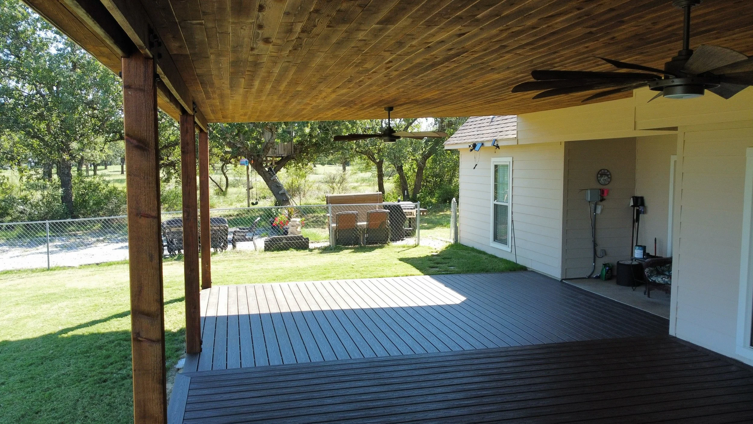 Covered patio with wooden ceiling and ceiling fans, overlooking a grassy backyard with trees, a chain-link fence, outdoor furniture, and a tree with a treehouse.