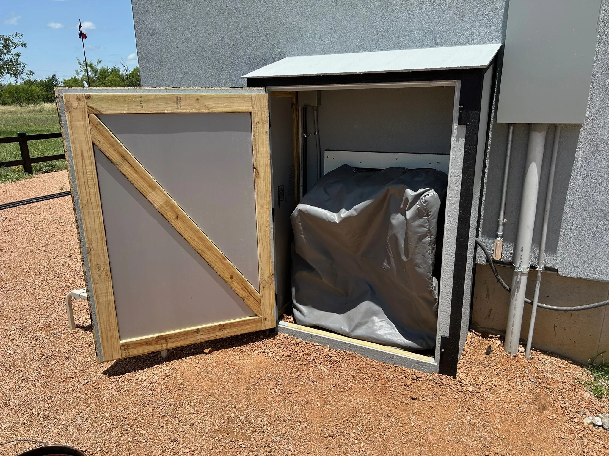 An outdoor scene showing partially assembled electrical or HVAC utility box with a wooden frame around an opening, attached to a building wall. Pipes and electrical conduits are visible on the right side. In the background, there are trees, a Texas f