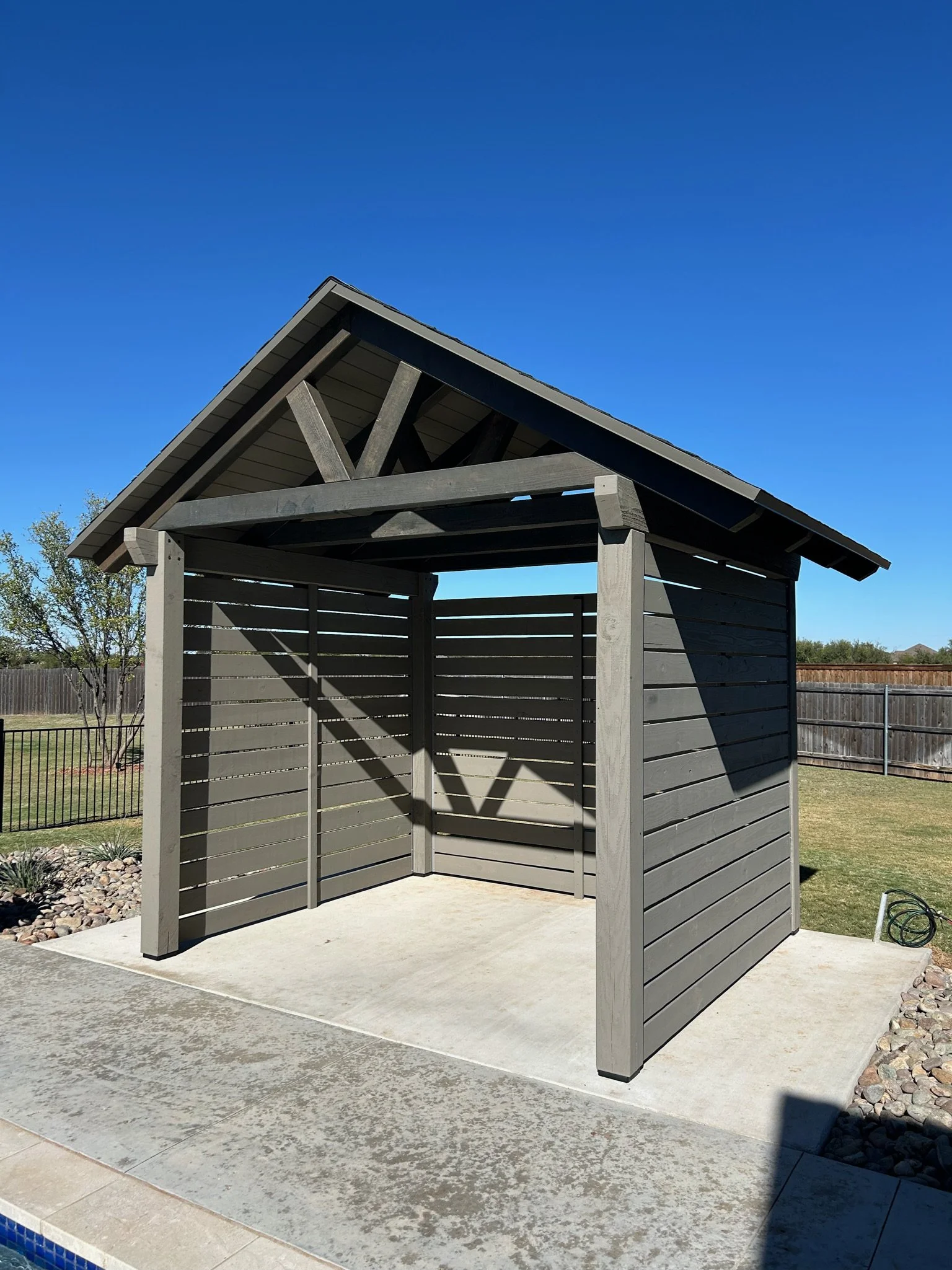 A small wooden outdoor cabana with an open front and slat walls, situated on a concrete patio near a swimming pool, with a clear blue sky overhead and a grassy yard with trees in the background.