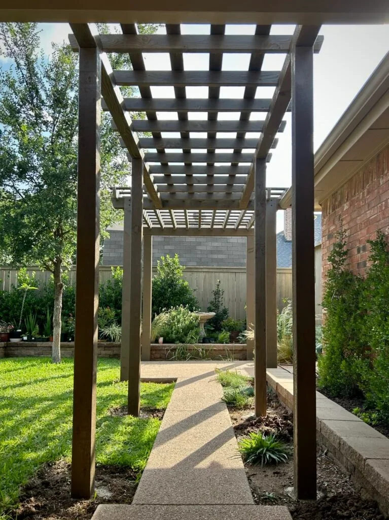 Wooden pergola over a concrete pathway in a backyard garden with trees, shrubs, and a brick house.