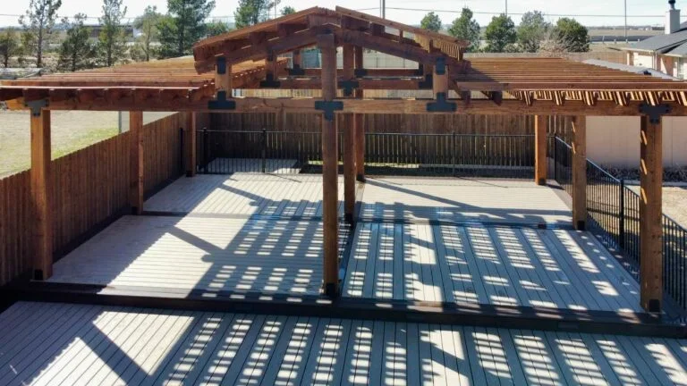 A wooden deck with a pergola, surrounded by a wooden fence and metal railing, casting patterned shadows on the surface.