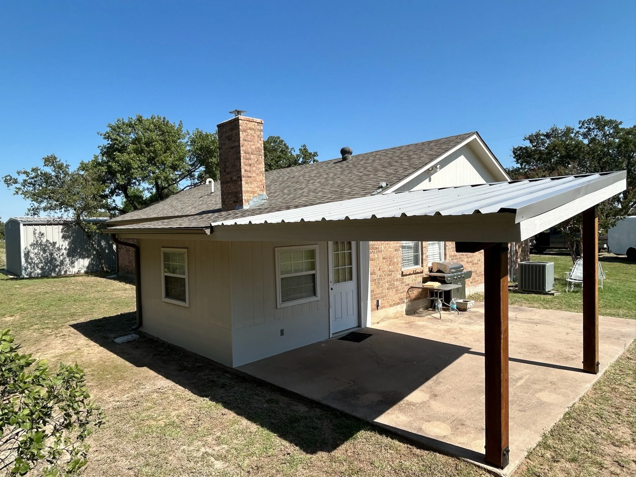 Backyard view of a house with a brick chimney, gray shingle roof, and white siding. There are trees and a clear blue sky in the background. Part of a patio cover with a white metal roof and wooden posts is visible in the foreground.