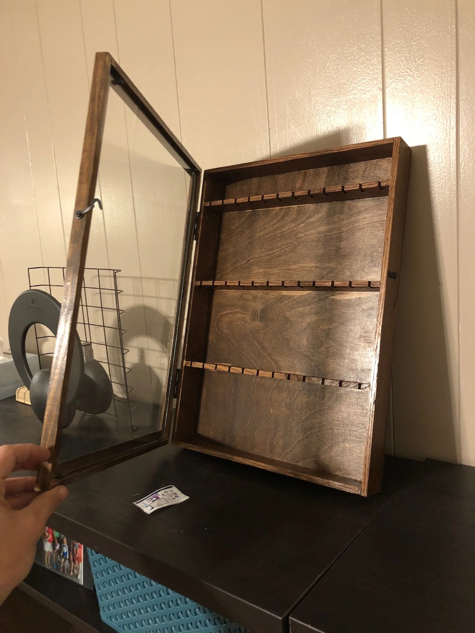 A person holding a wooden and glass display case with a top lid open, showing empty shelves inside. There is a blue basket and some hanging objects visible below the table.