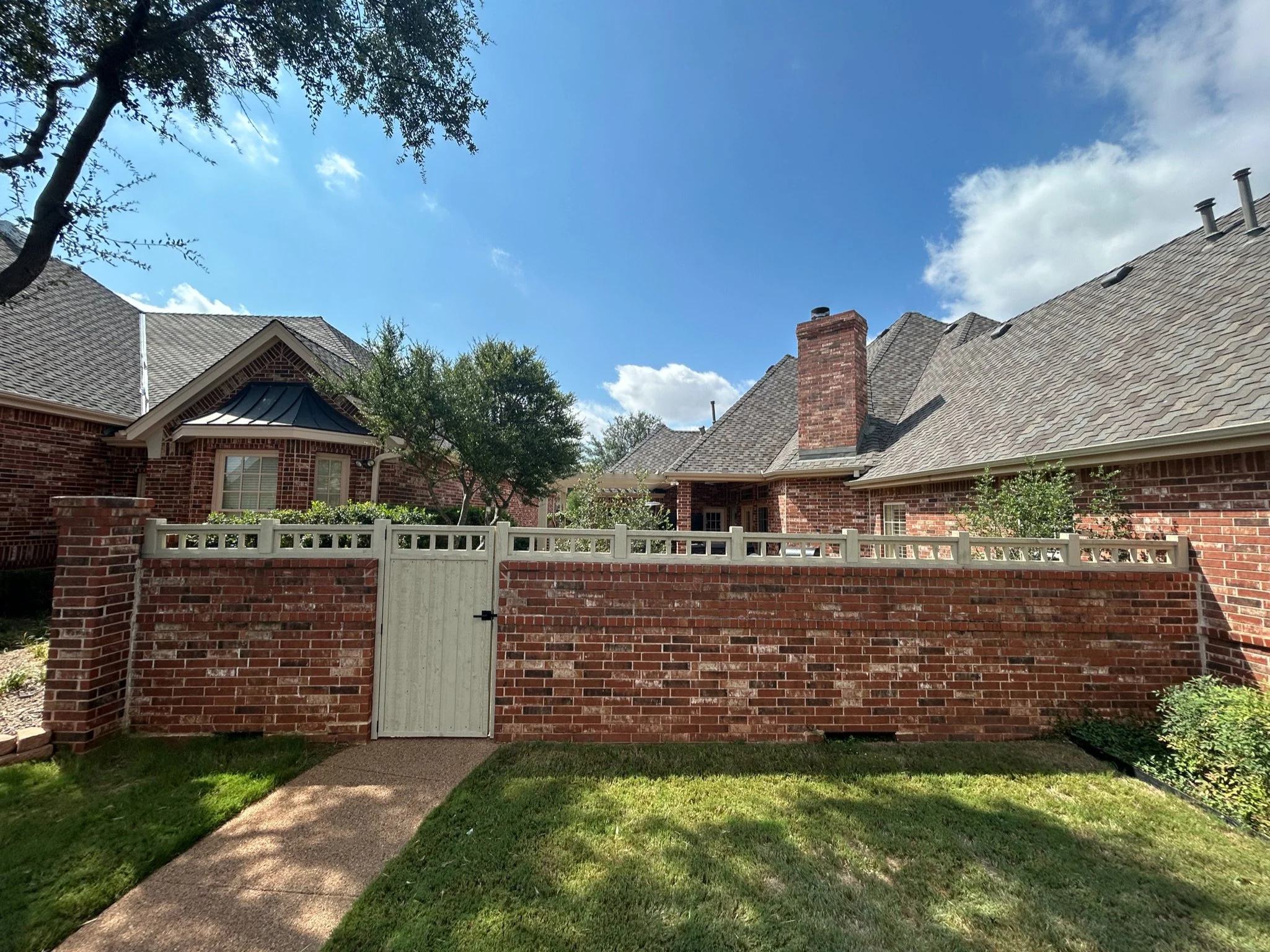 Red brick house with a tall chimney, gray shingle roof, and a white wooden gate in front of a brick fence, green lawn, trees, and a bright blue sky with some white clouds.