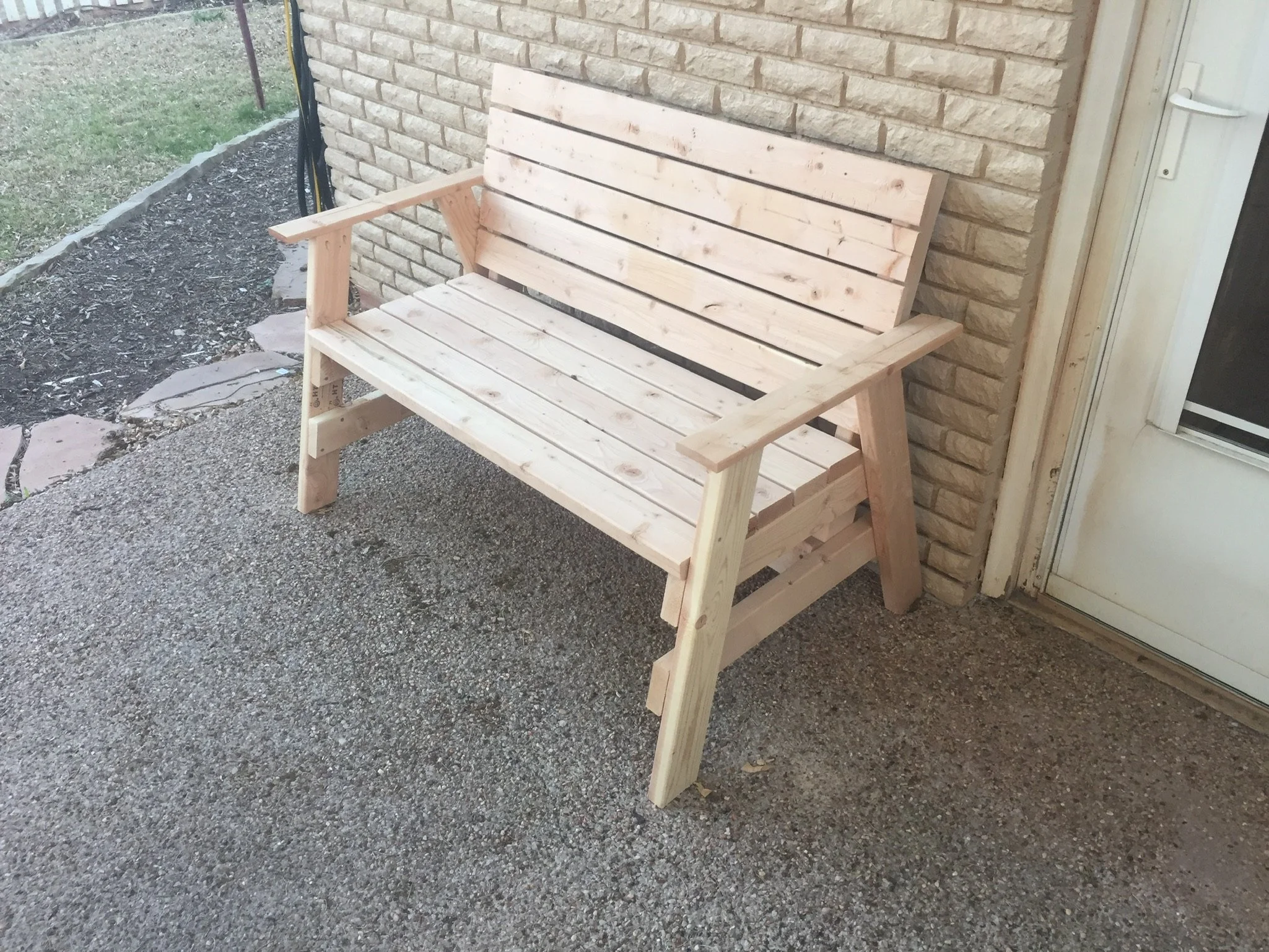 Unfinished wooden bench placed outdoors near a house wall with brick siding, next to a glass door with a screen. The ground is a mixture of dirt and gravel.