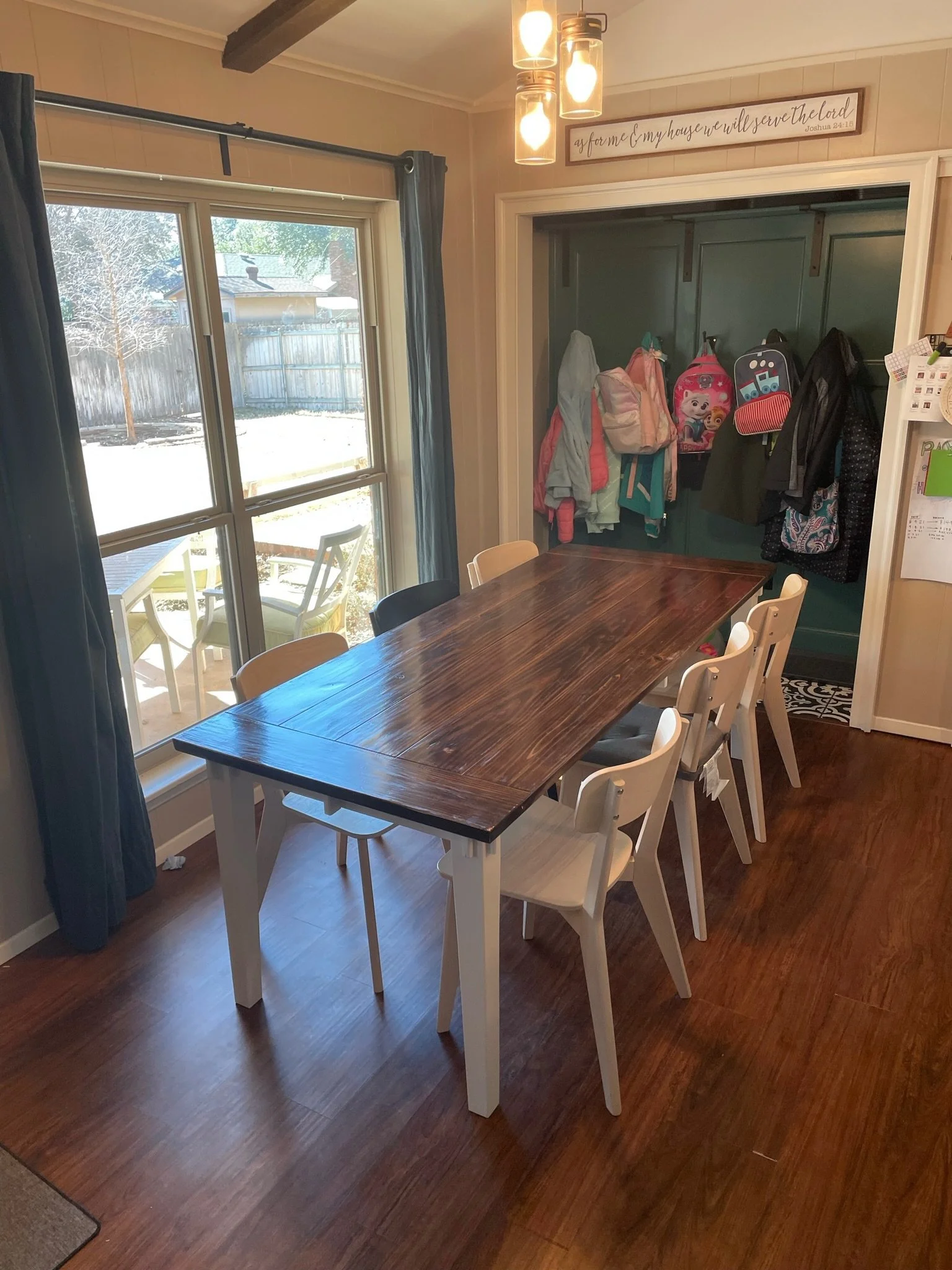 A dining room with a wooden table surrounded by white chairs, a large window with blue curtains, a sliding glass door outside leading to a patio with yellow chairs, and a coat rack with jackets and backpacks. There is a wooden sign with a printed Bib