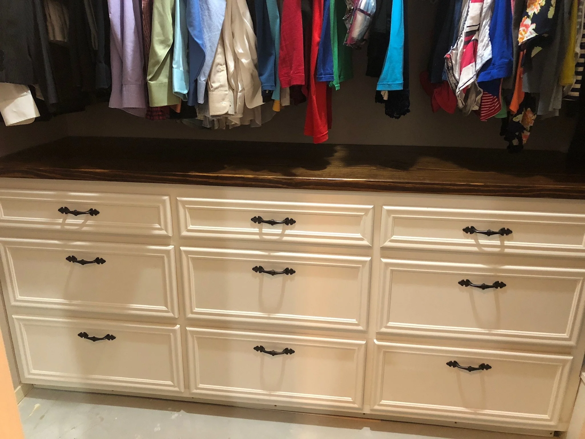 A white dresser with dark handles and a wooden top, partially visible in the lower part of the image, with a row of various colorful shirts hanging above on a rack.