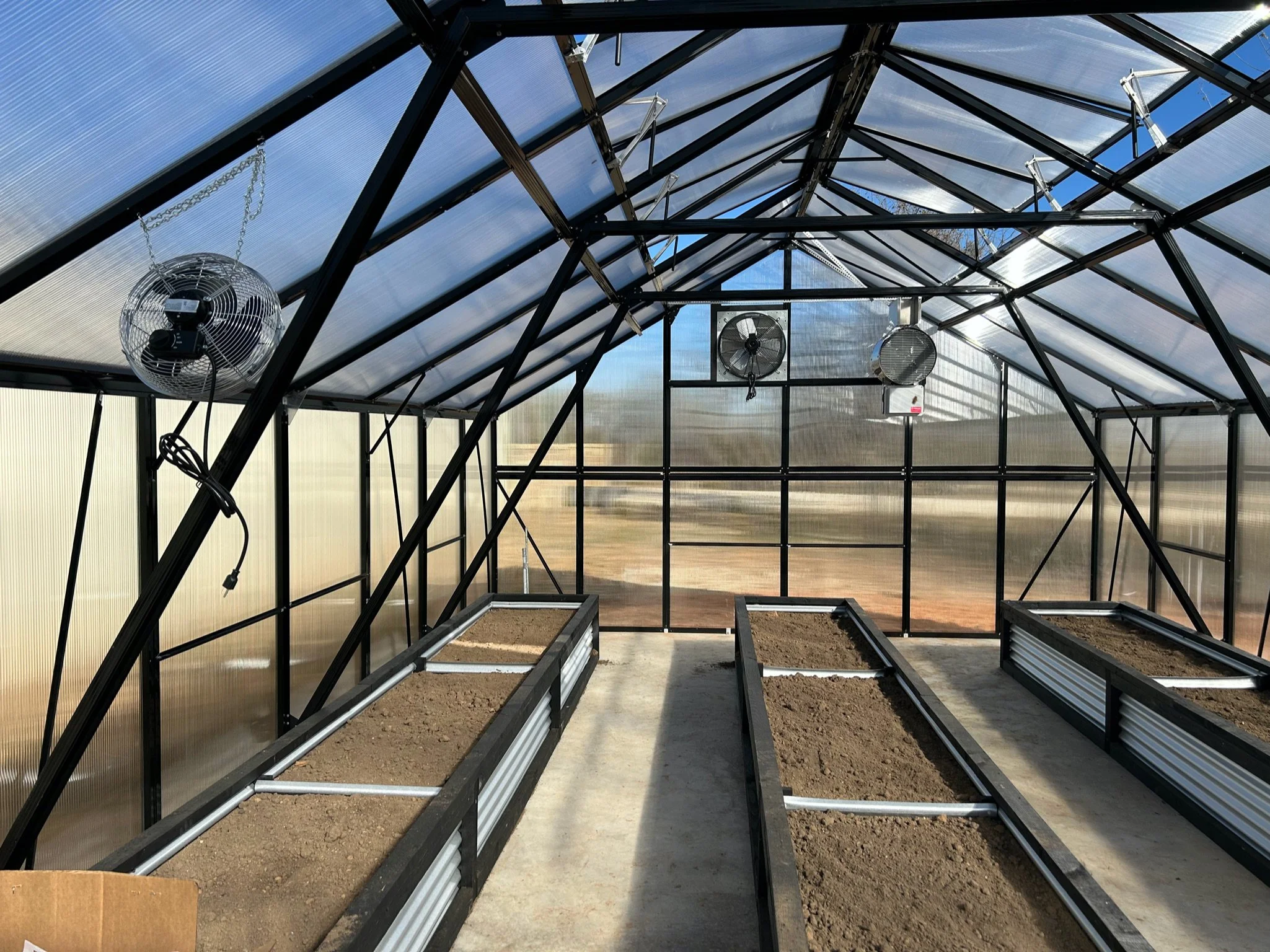 Inside a greenhouse with a metal frame structure and transparent roof panels, equipped with hanging fans, and empty planting beds with soil.
