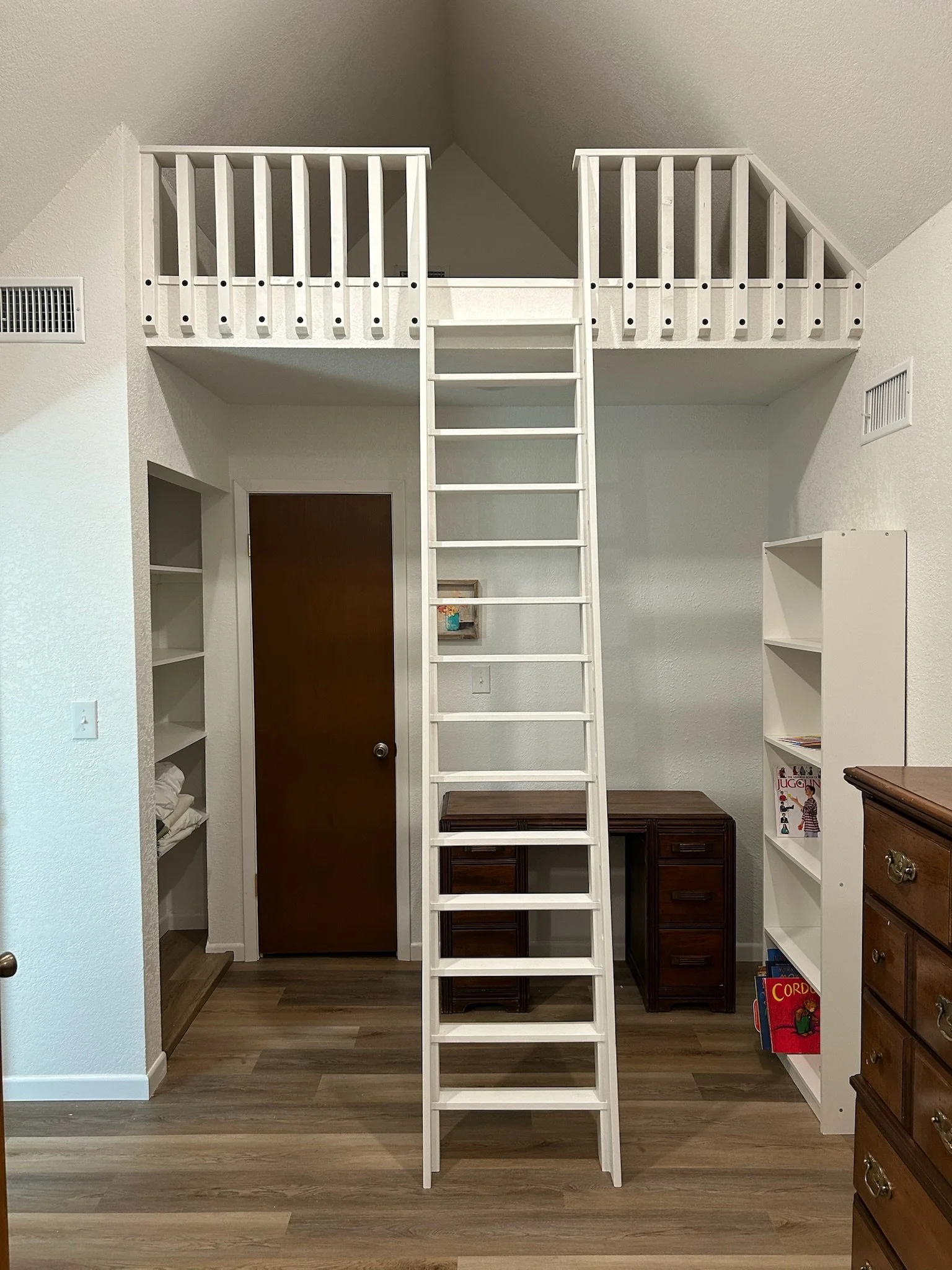 A white wooden ladder in the center of a room with ceiling sloped walls. Behind the ladder is a dark wooden door, and to the right, a white bookshelf with books and magazines. The room has light-colored walls and wood flooring.