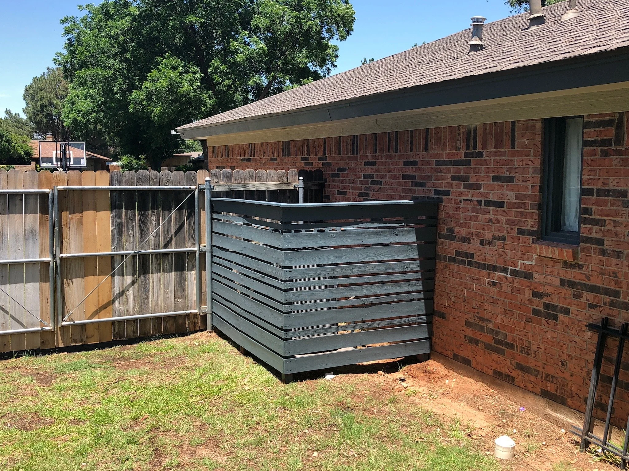 Backyard with wooden fence, brick house, raised wooden deck, ladder, trees, and a basketball hoop in the distance.