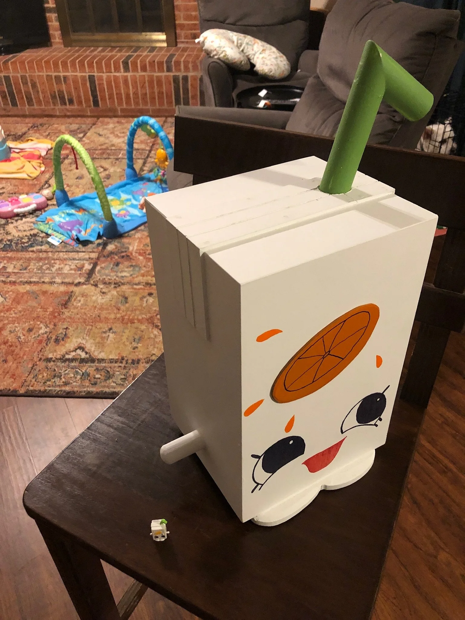 A white wooden toy cart with a painted panda face on the front, featuring orange and black details, on a dark wooden table. In the background, there are a gray couch with a patterned pillow, a brick fireplace, and children's toys on a colorful rug.