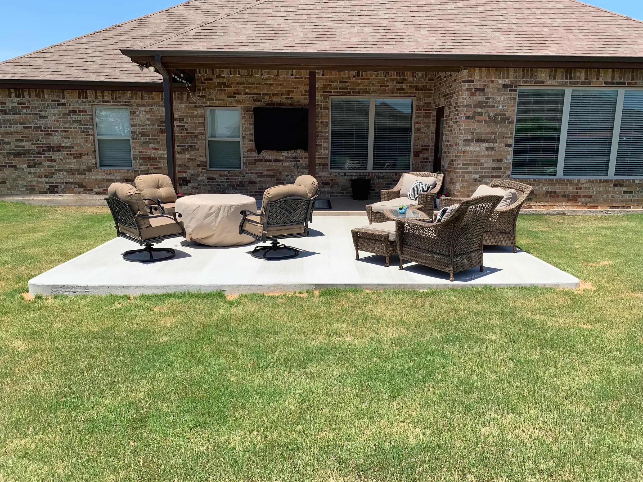 A backyard patio with outdoor furniture including chairs, a table, and cushions, with a brick house wall and windows in the background.