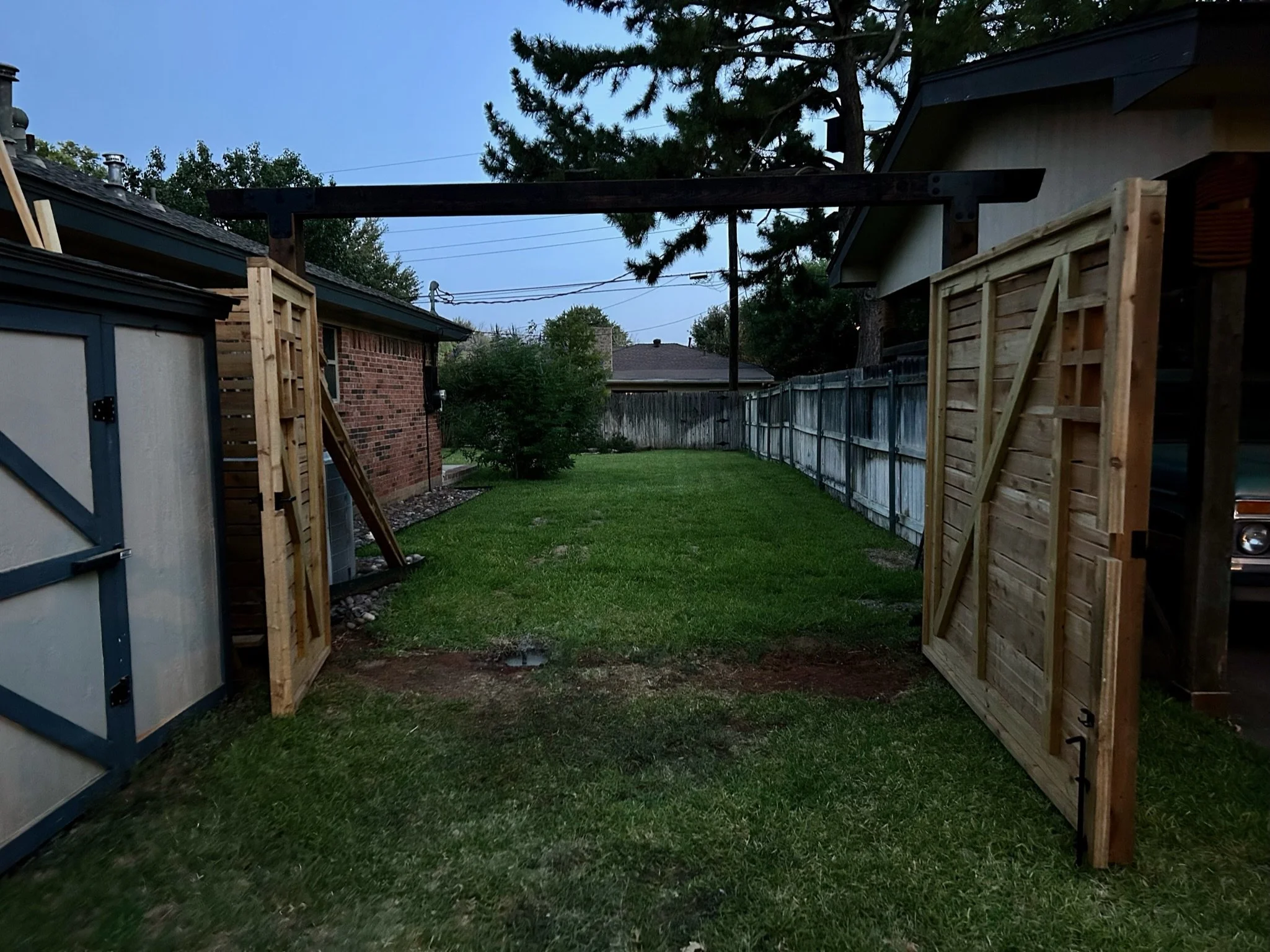 Backyard with wooden structures and open gate, green grass, trees, and houses in the background under a cloudy sky.