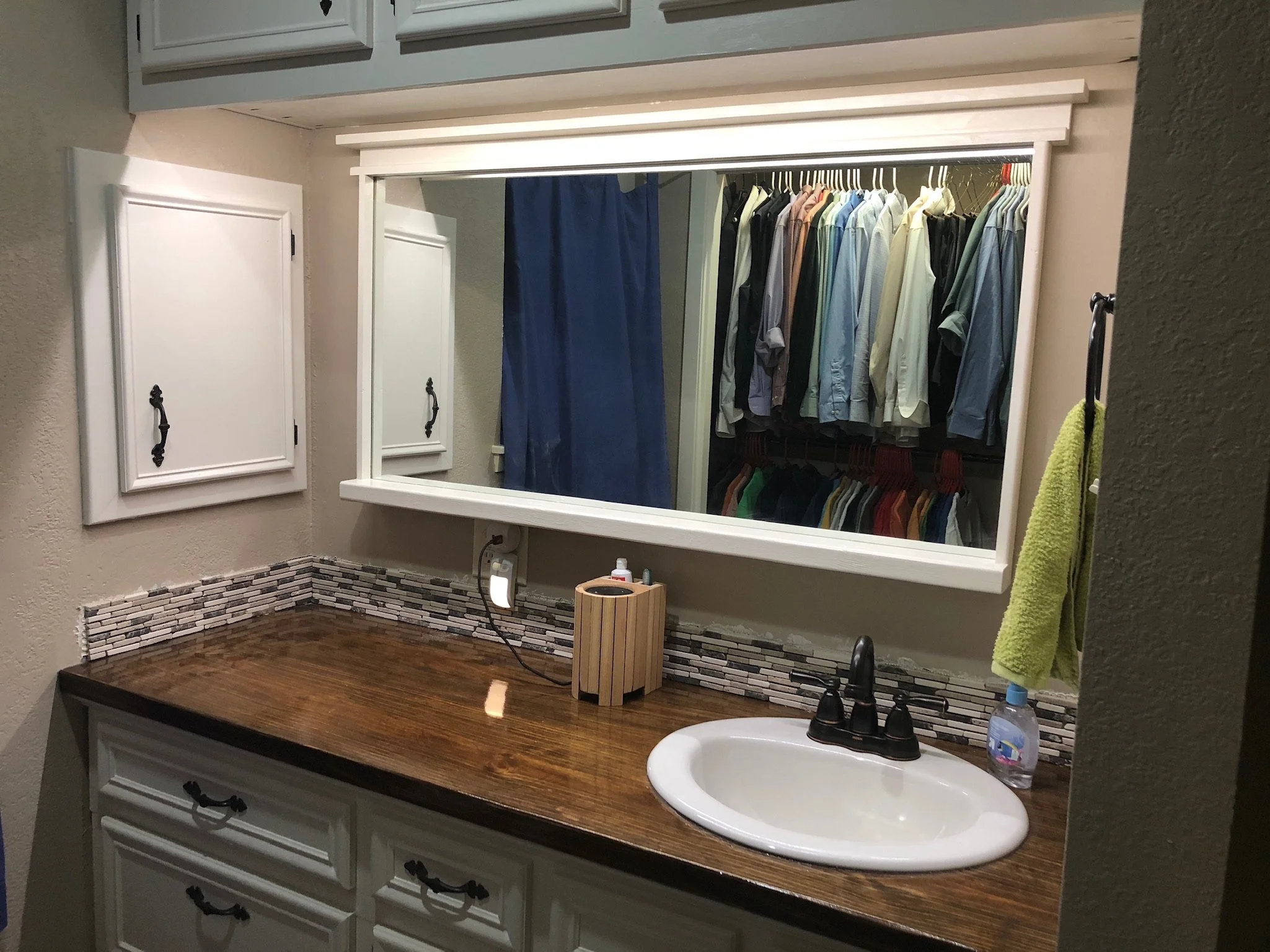 Bathroom vanity with a wooden countertop, white cabinet doors, and a large mirror. Clothing is visible through the mirror reflection, hung in a closet. Items on the countertop include a small wooden box, a bottle of hand soap, and a plastic water bot